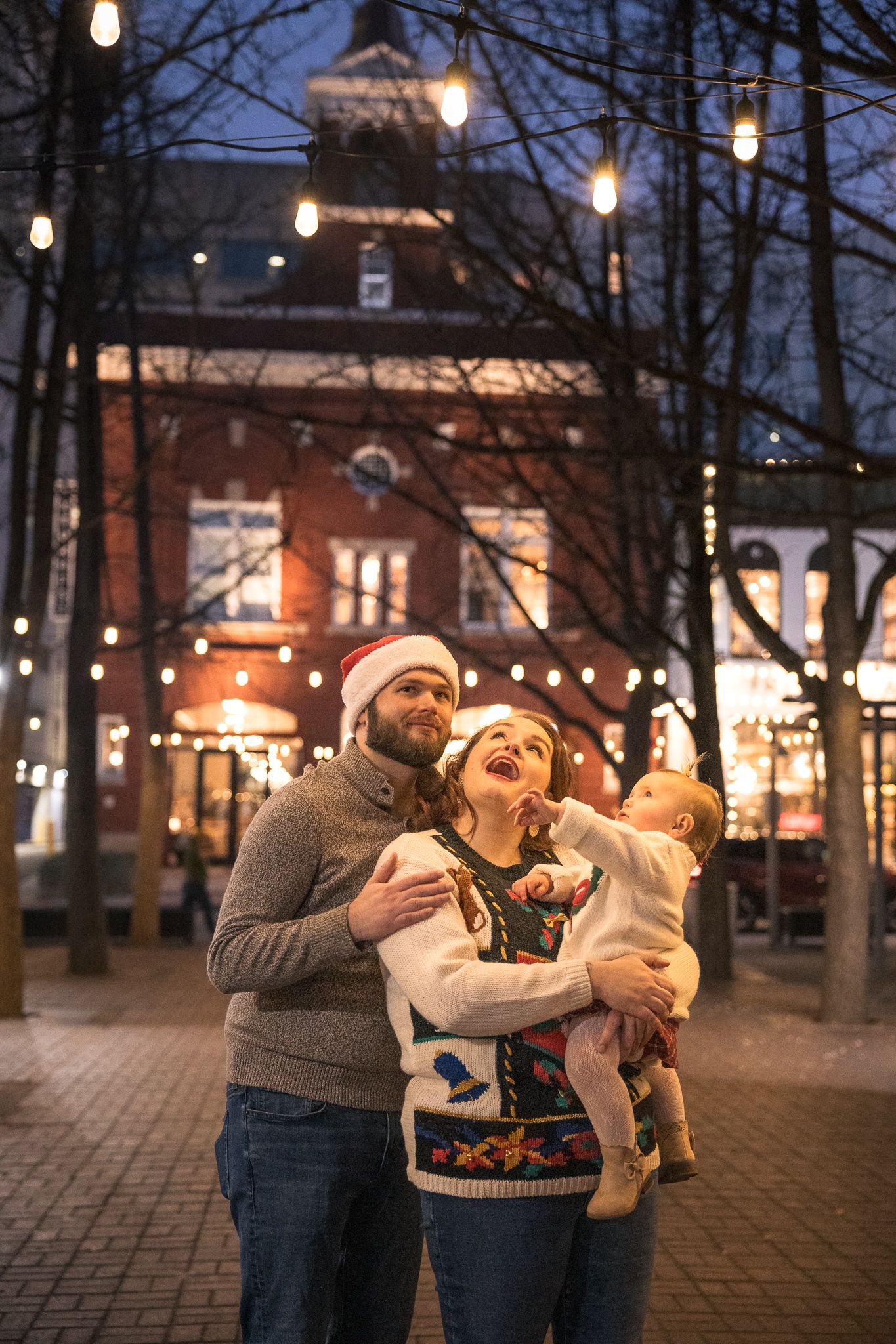 Happy family with baby celebrating Christmas outdoors at night with festive lights and decorations.