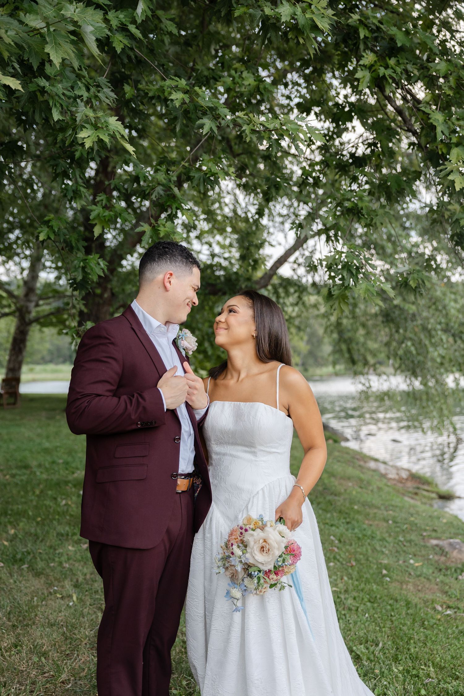 Bride and groom sharing a loving gaze under green trees near a pond, groom in burgundy suit with boutonniere.