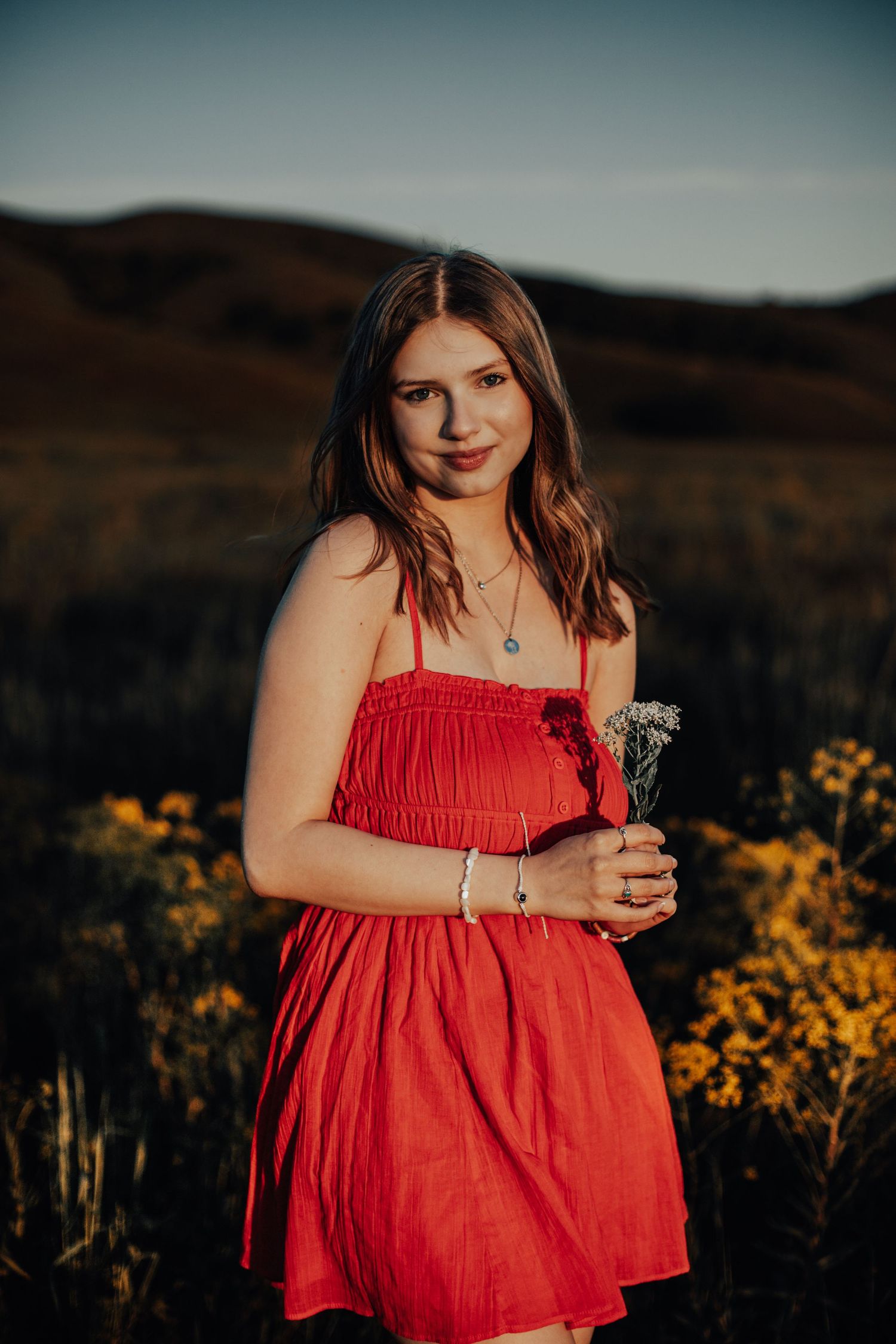 Smiling teenage girl in red dress holding wildflowers in a field of yellow flowers at dusk with hills behind.
