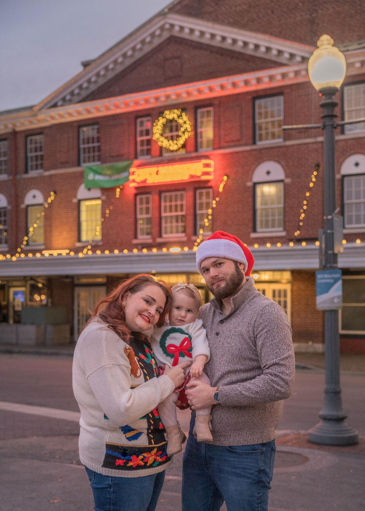 Family of three poses outdoors at Christmas, parents holding toddler in Santa hat before festive holiday decorated building.