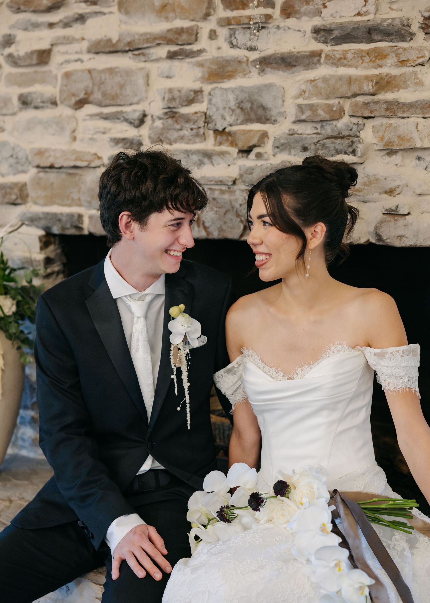A young couple in wedding attire smile at each other in front of a stone fireplace, bride holding white flowers.