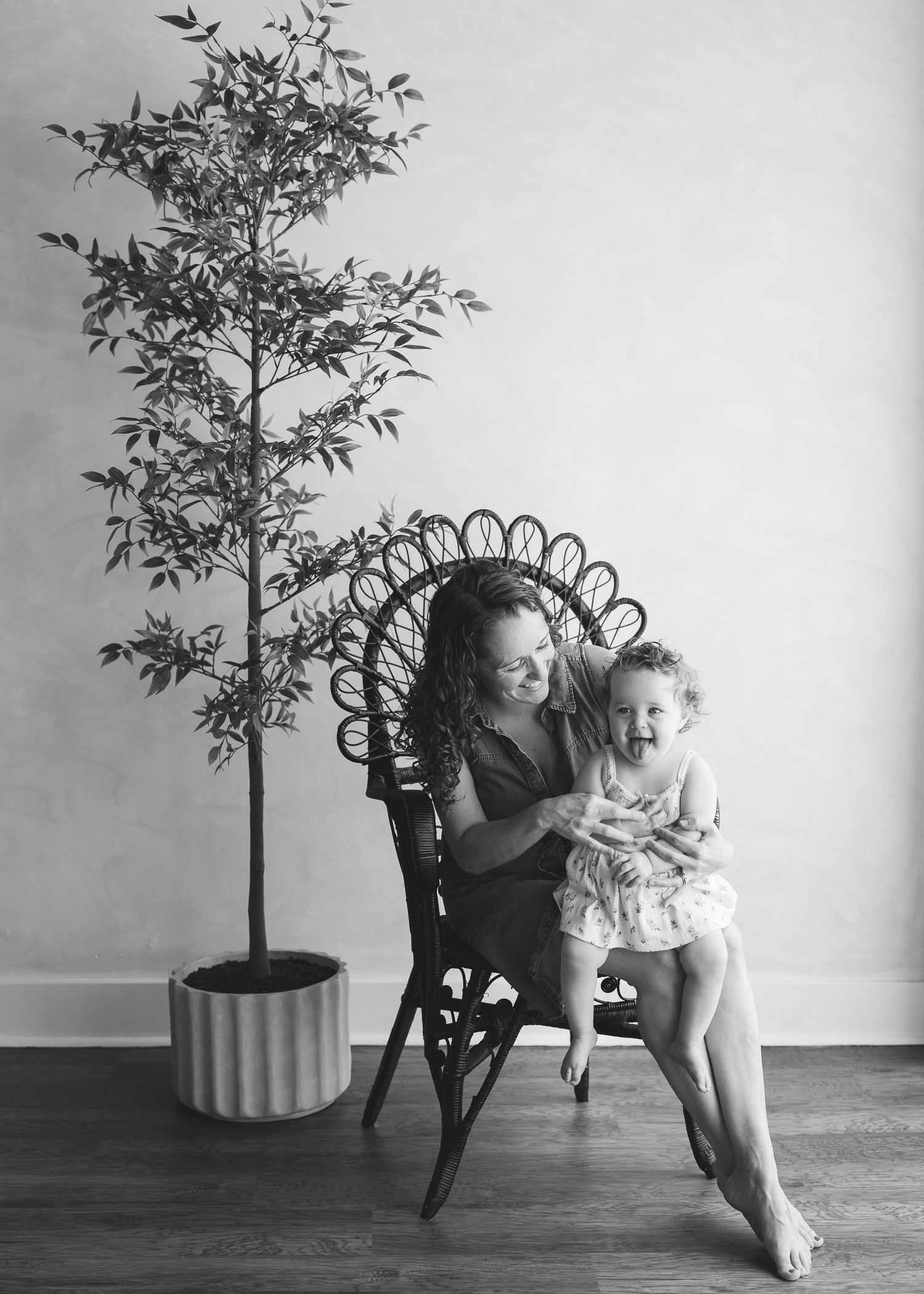 Black and white photo of a mother and toddler laughing together in a wicker chair beside a potted tree.
