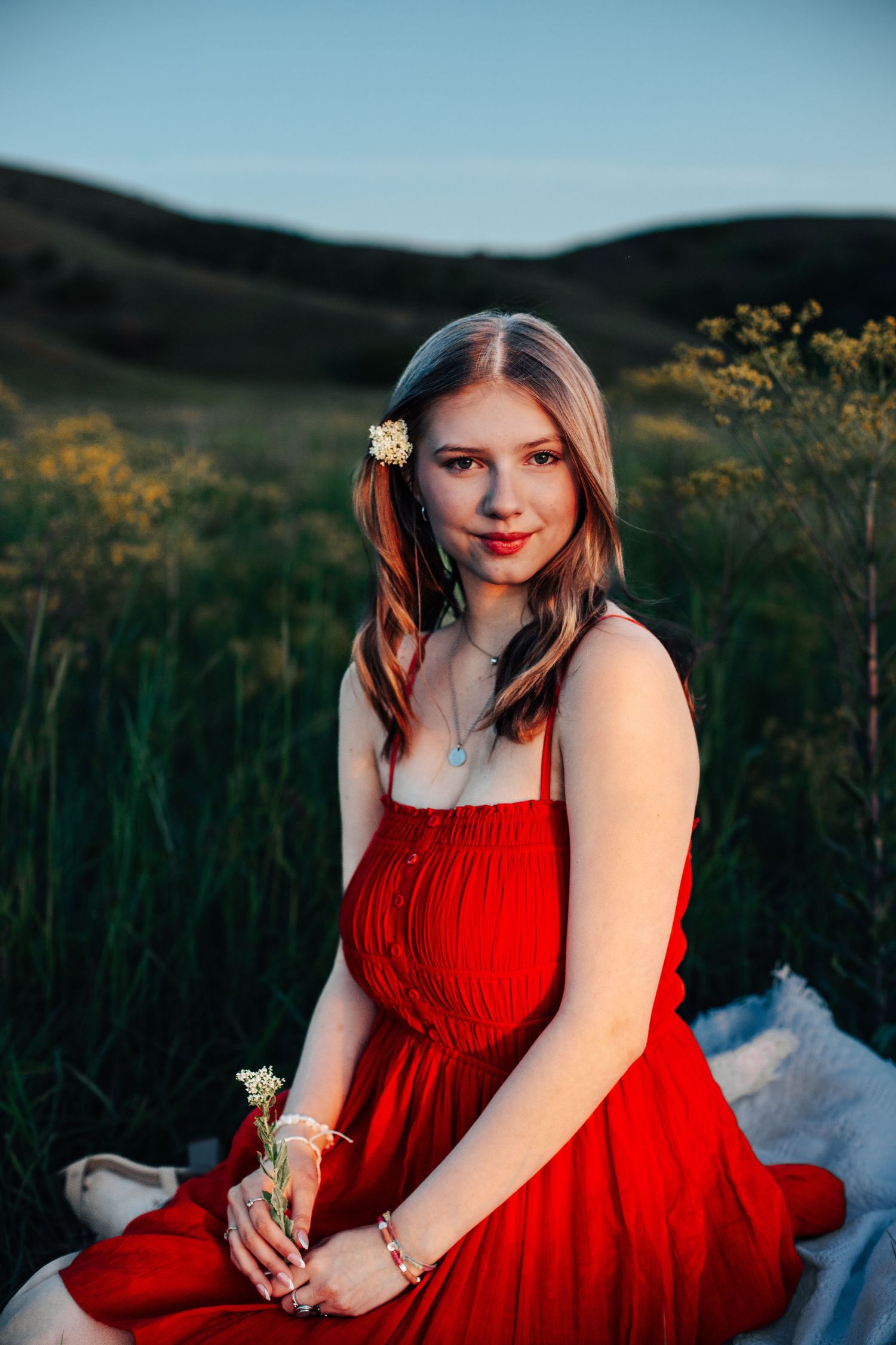Young woman in red dress sitting in a grassy field holding white wildflowers with a flower tucked in her hair.