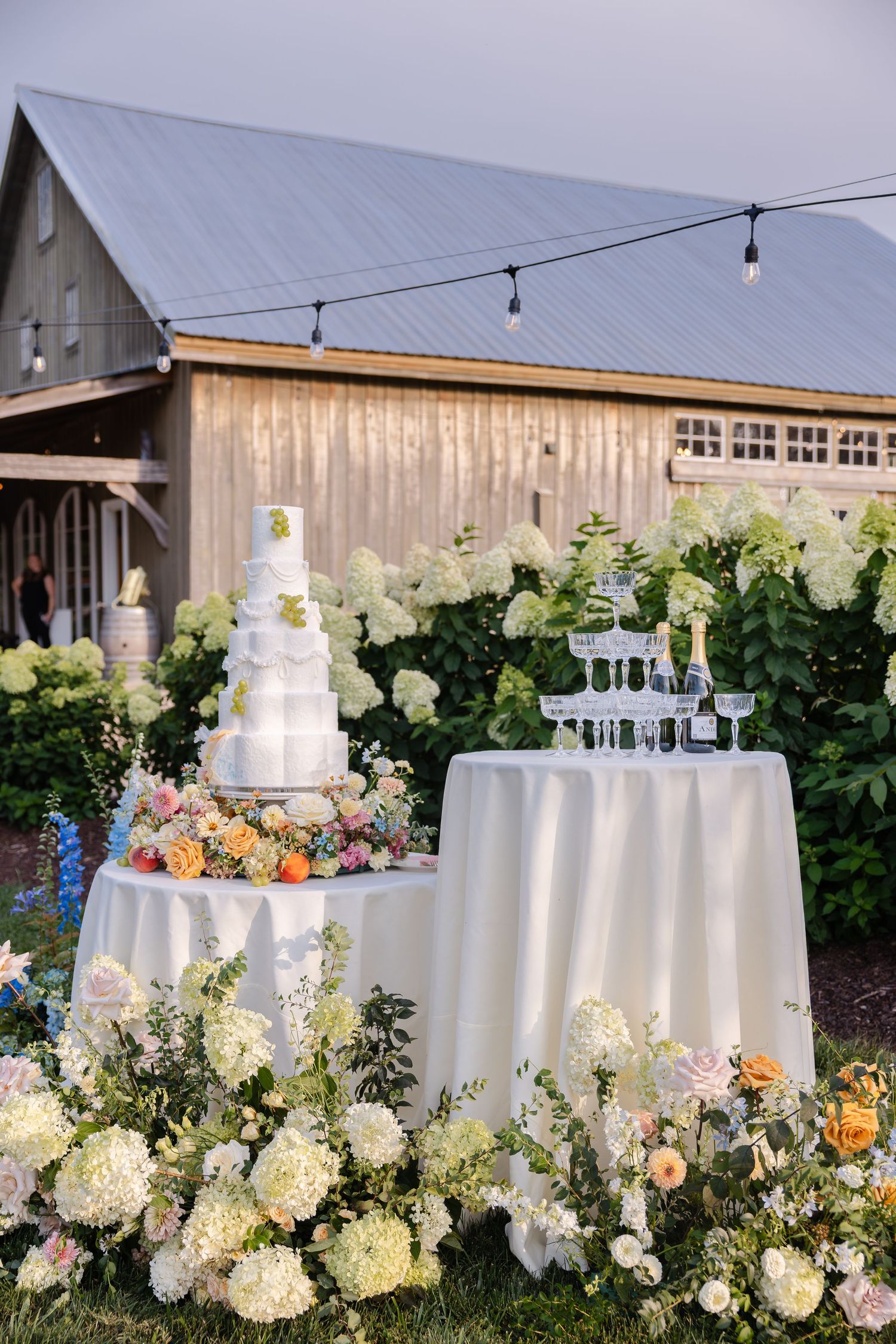 Outdoor wedding cake table with white floral arrangements and champagne tower beside a rustic barn venue.
