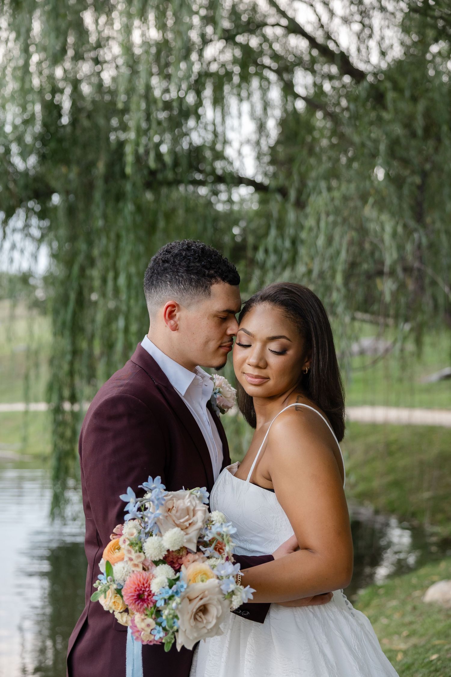 Groom in burgundy suit tenderly embracing bride holding a pastel bouquet near a willow tree and pond.