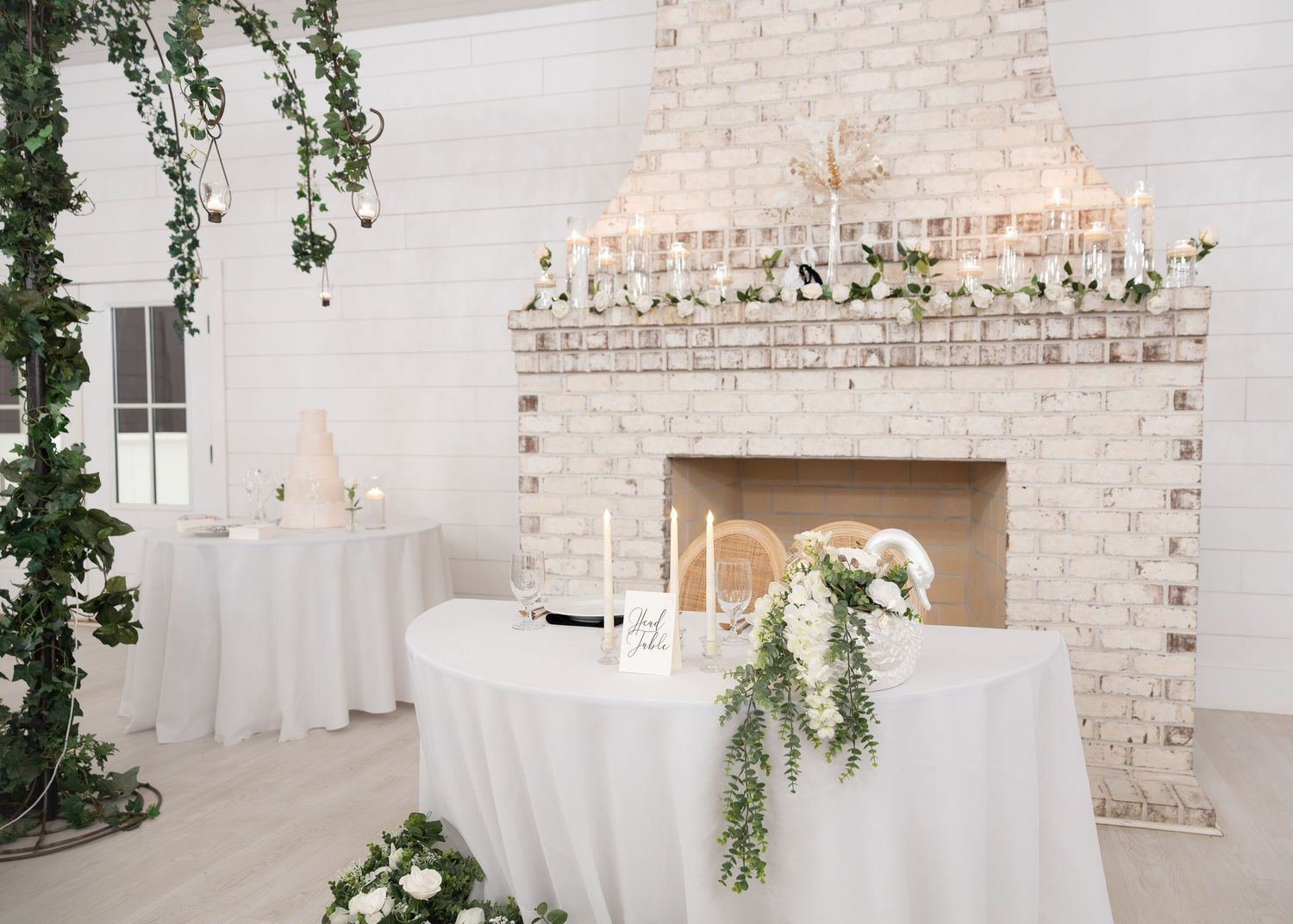 Elegant wedding reception setup with white draped tables, floral greenery, candles, and a whitewashed brick fireplace backdrop.