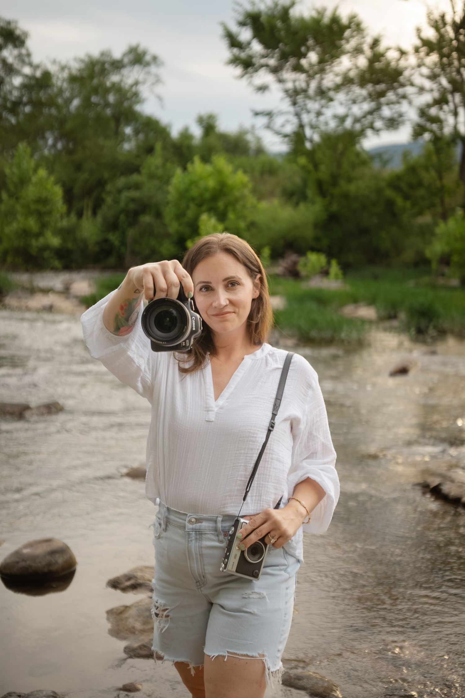 Female photographer points DSLR camera toward viewer while standing at a scenic rocky riverbank.