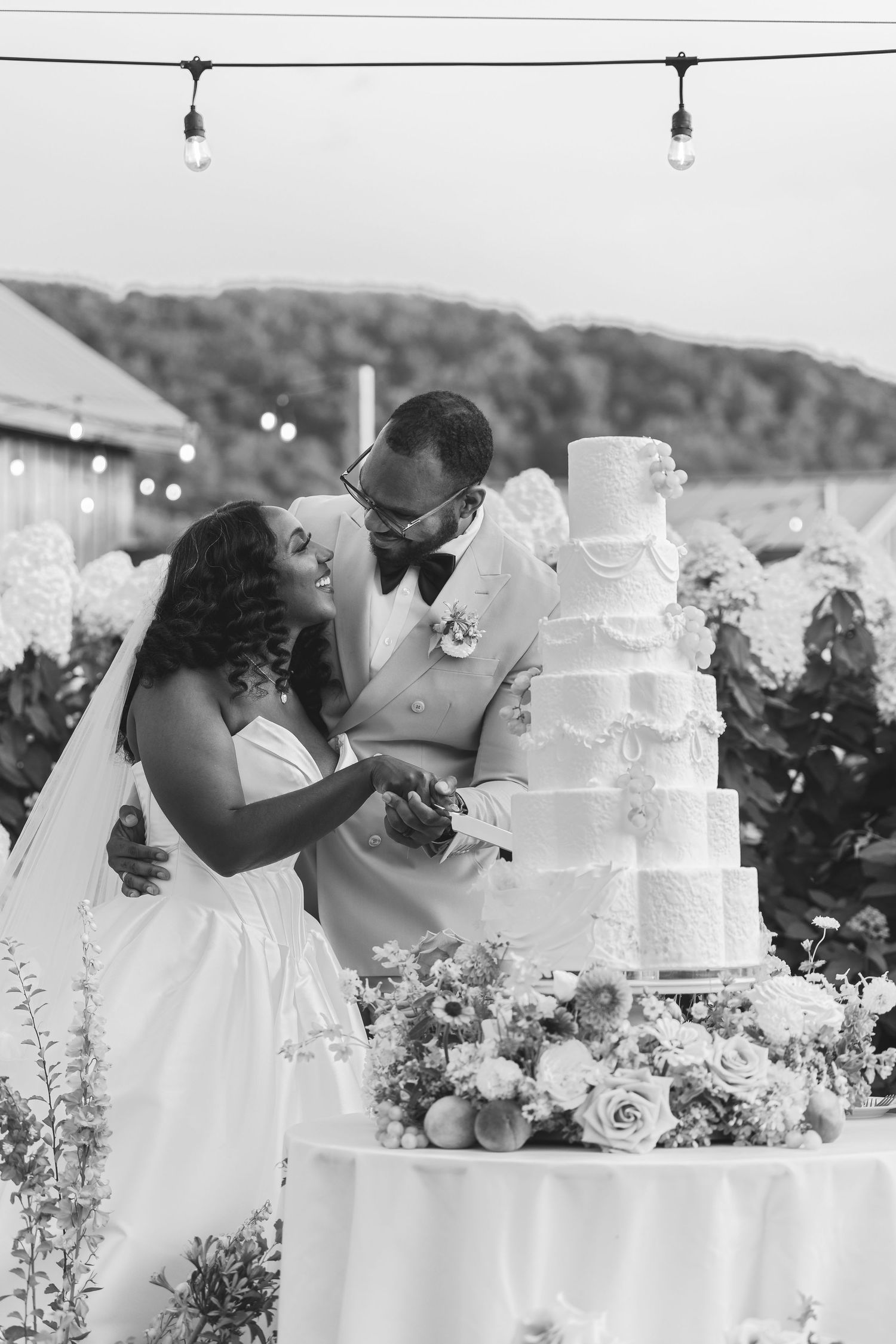 Black and white photo of bride and groom cutting a tall five-tier wedding cake surrounded by hydrangeas and roses.
