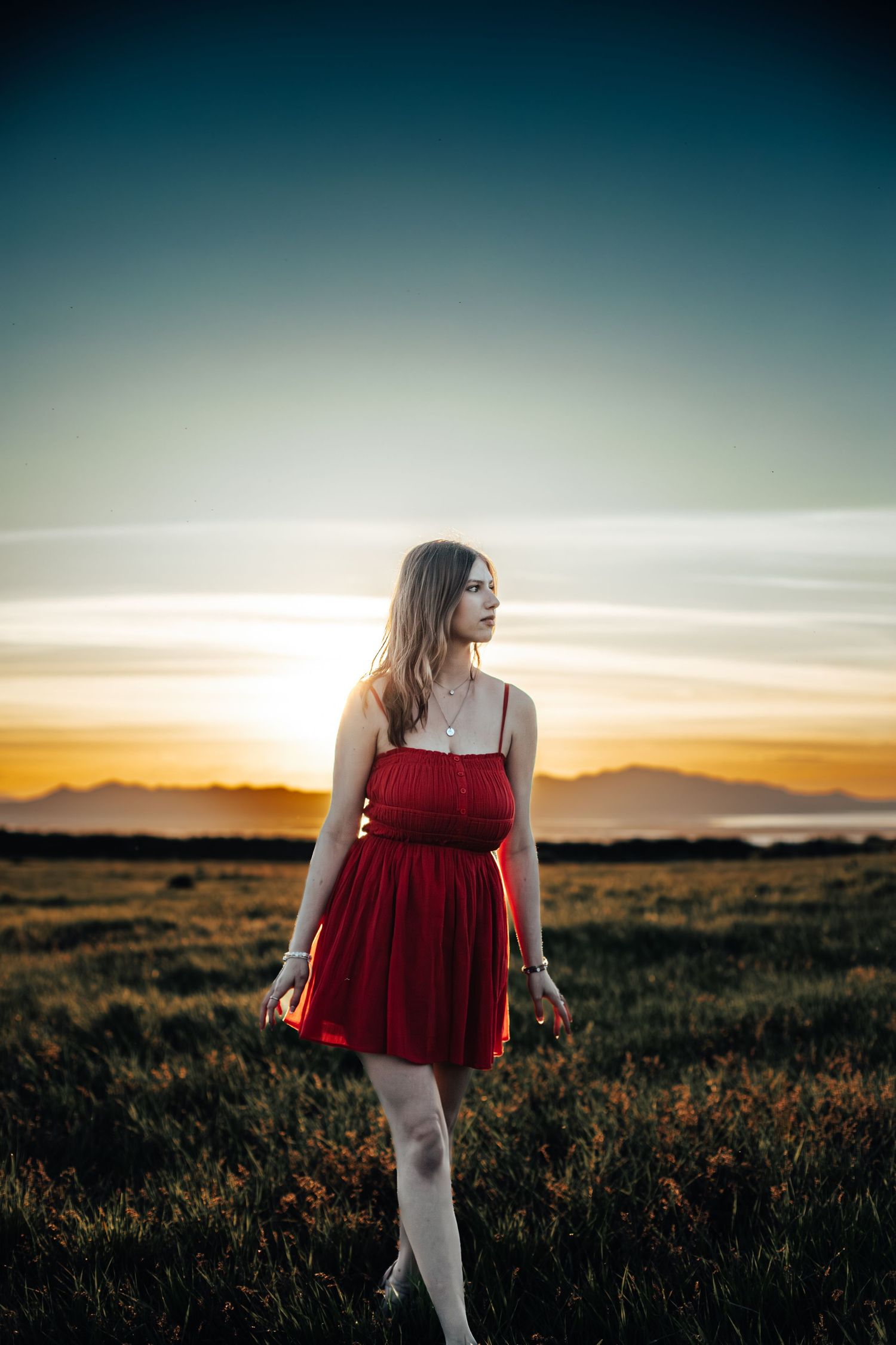 Young woman in red sundress standing in a field at golden hour with mountains and sunset sky behind her.