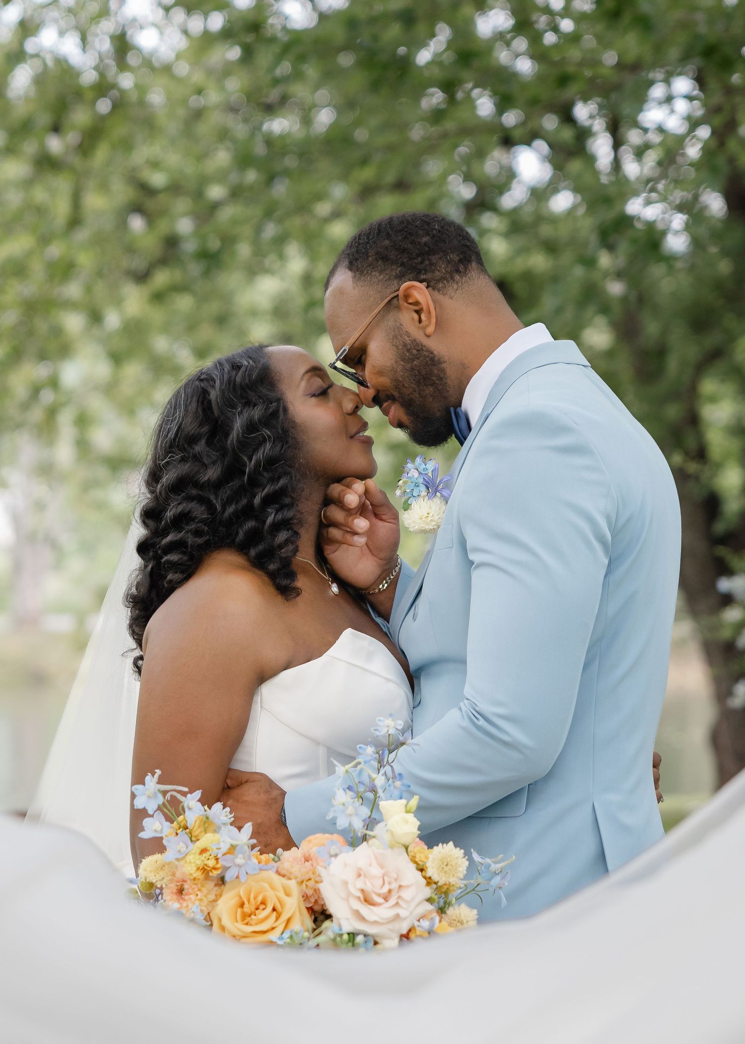 Black couple in wedding attire share a romantic moment, bride holding colorful bouquet, groom in light blue suit.