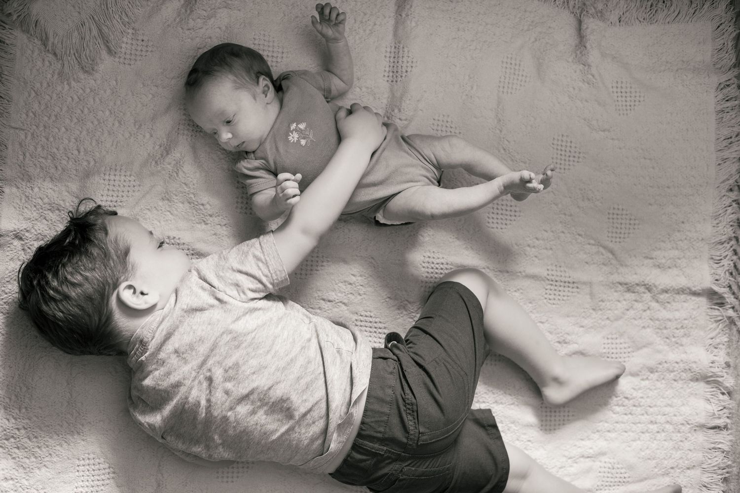 Black and white photo of an older child lying down holding a newborn baby on a soft blanket.
