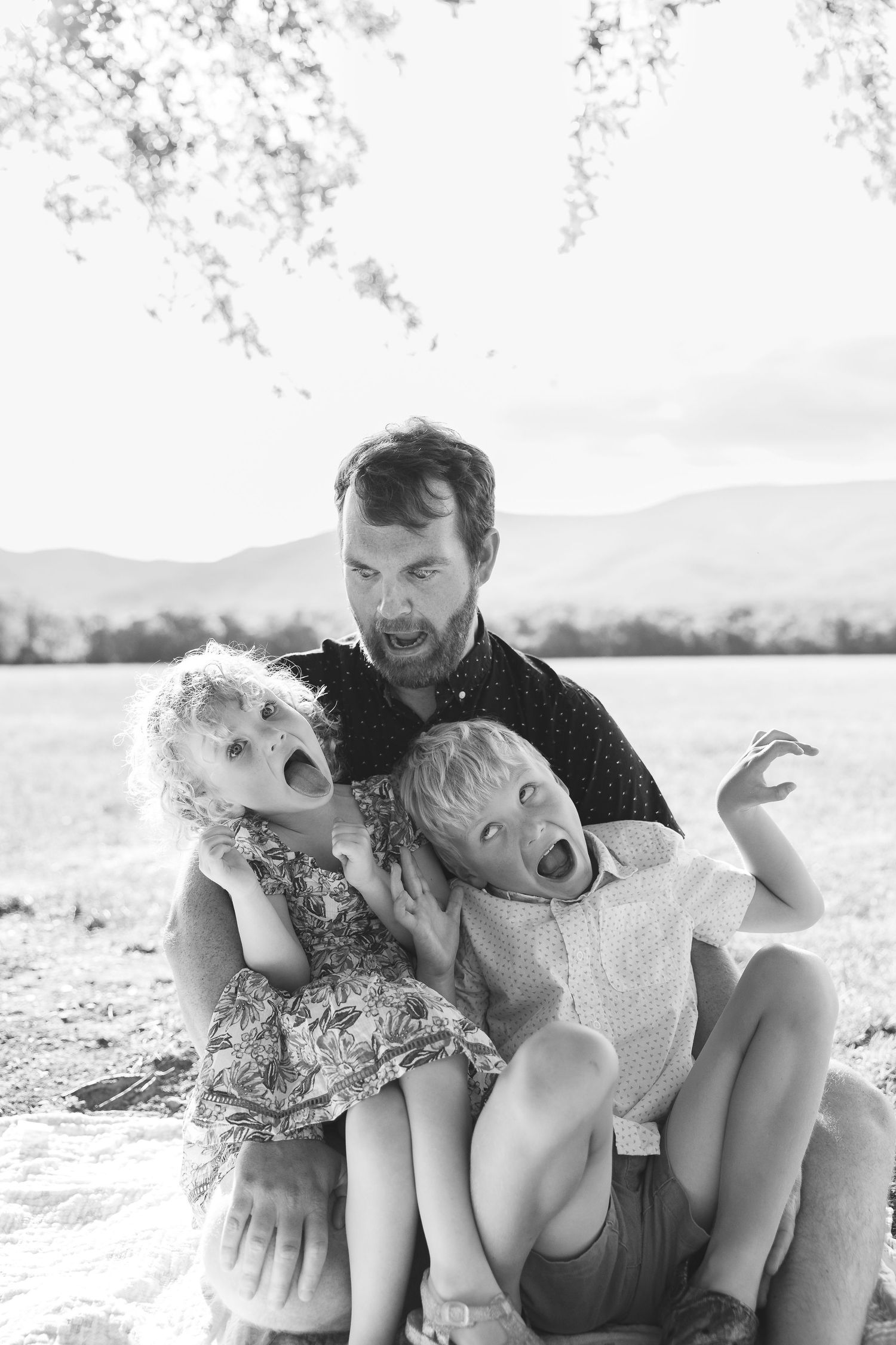 Black and white photo of a father and two young children making silly screaming faces outdoors near mountains.