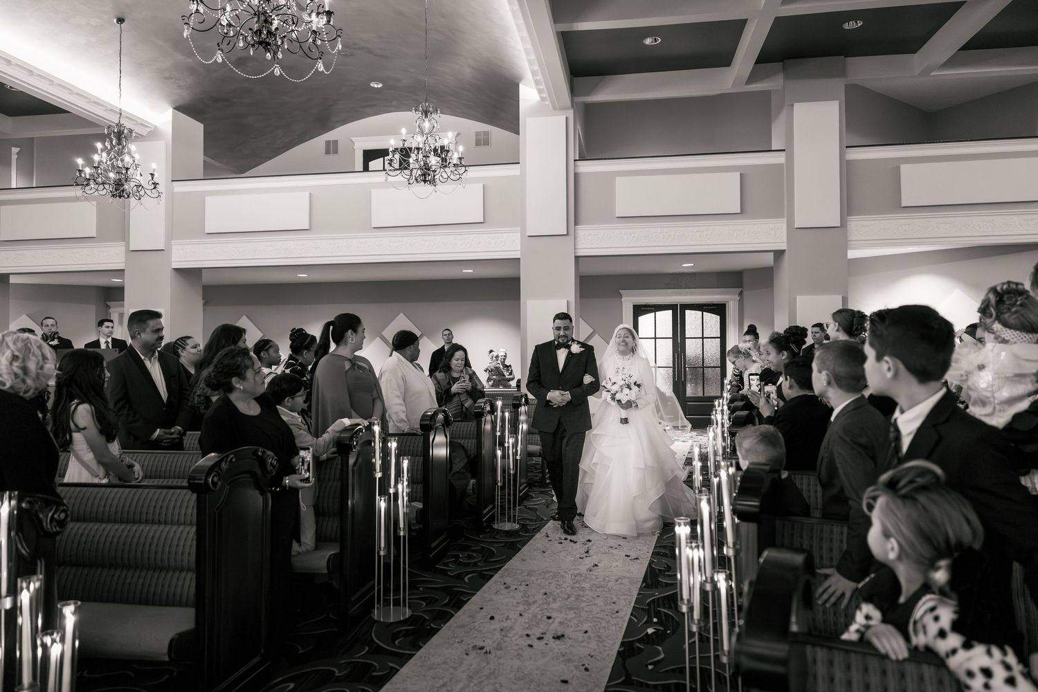 Black and white wedding ceremony photo of bride walking down the aisle with guests seated in elegant venue.
