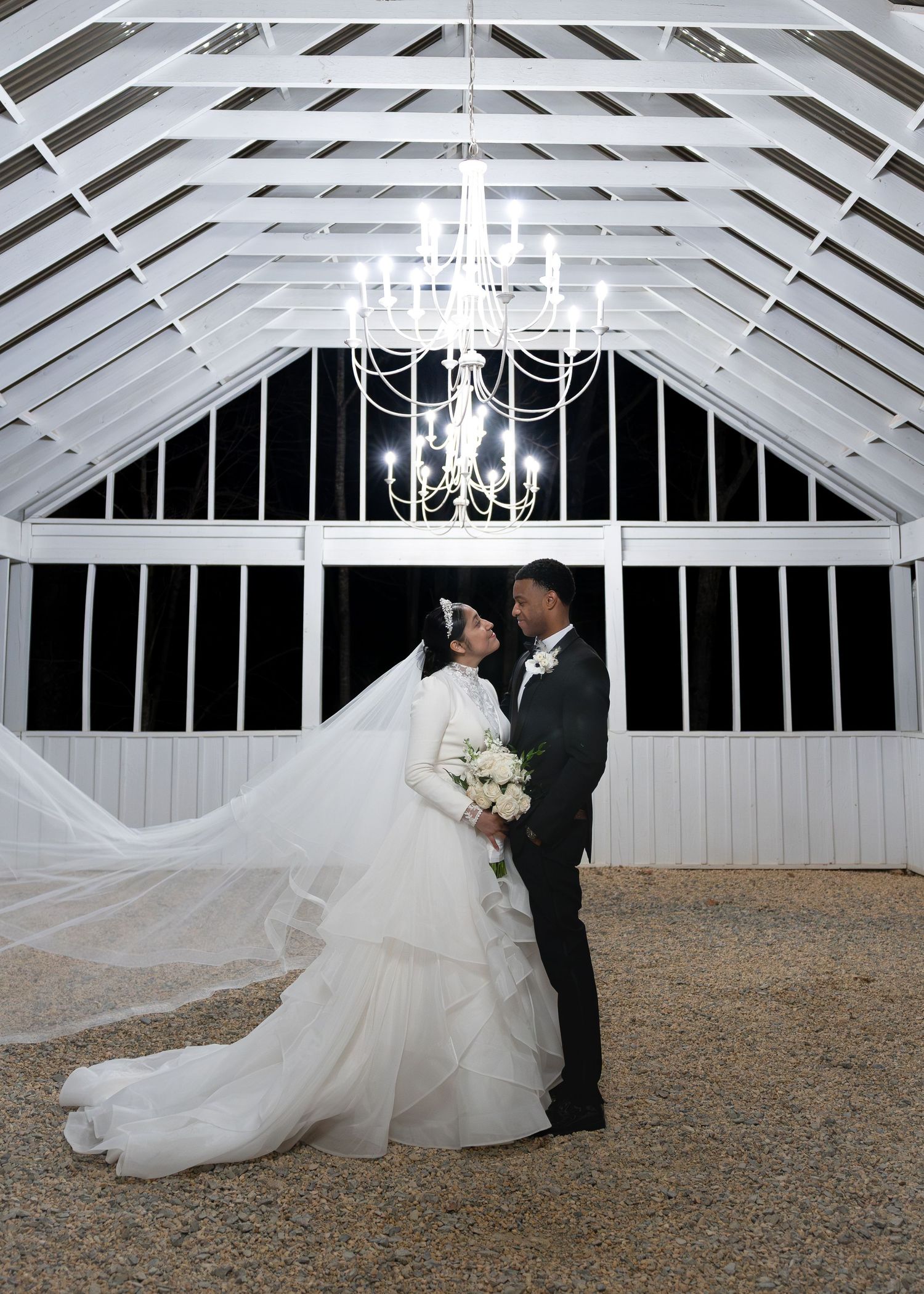 Romantic newlyweds kissing under chandelier in white barn venue, bride's veil flowing dramatically.