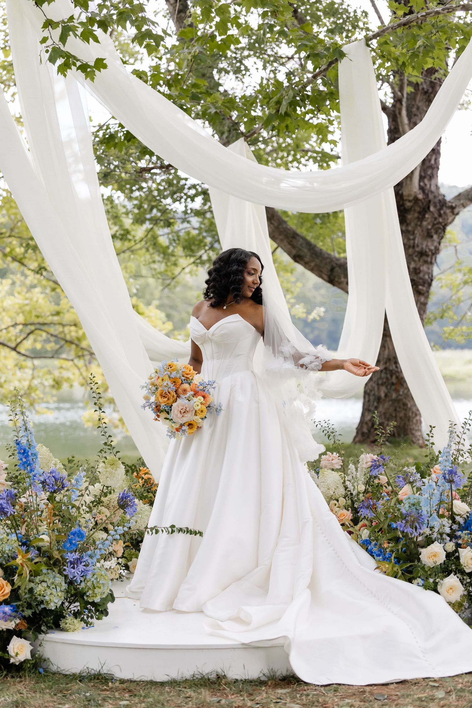Elegant bride in off-shoulder white gown with veil posing under white drapes surrounded by flowers.