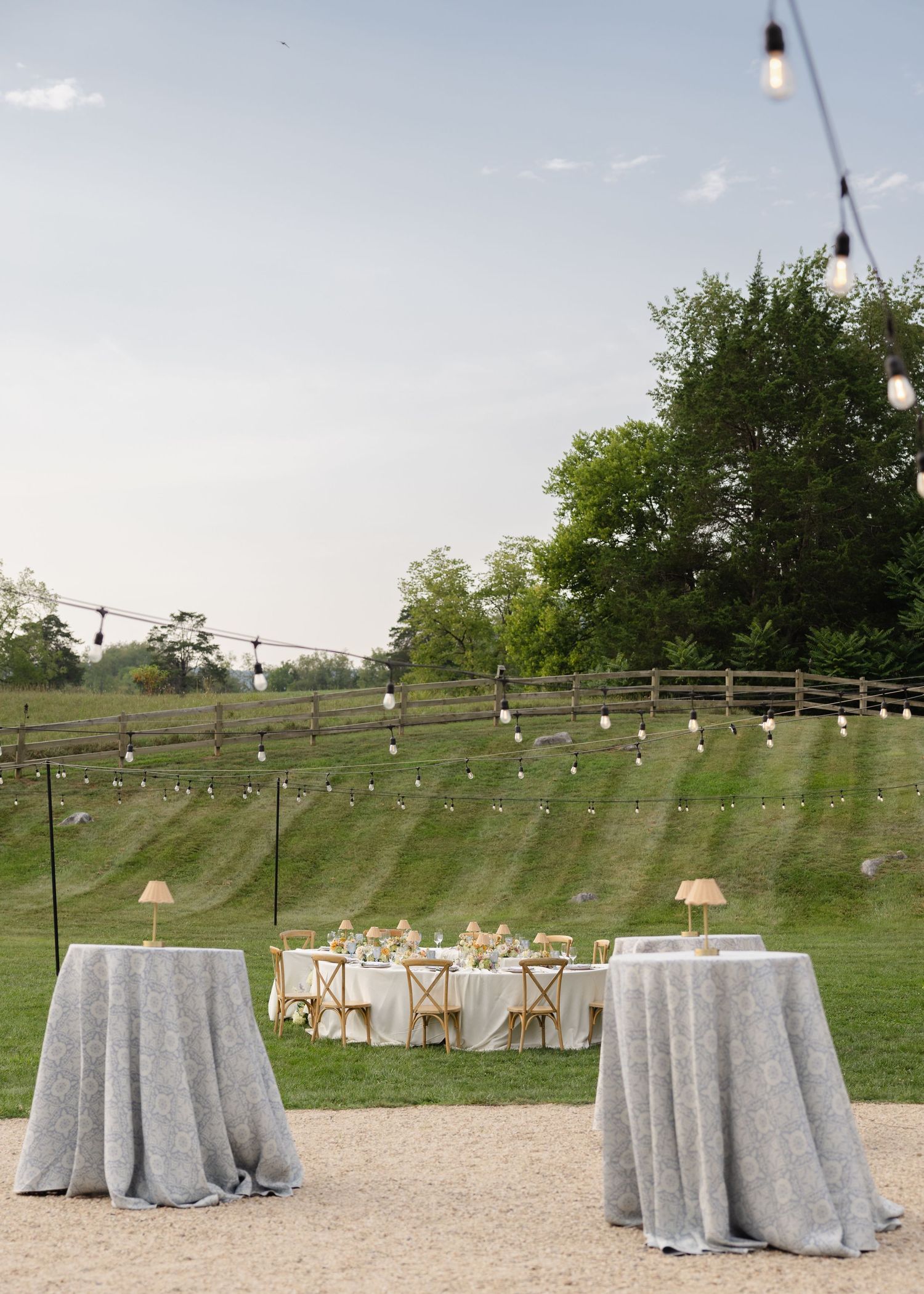 Outdoor wedding reception setup at dusk with draped cocktail tables, string lights, and a rustic wooden fence backdrop.