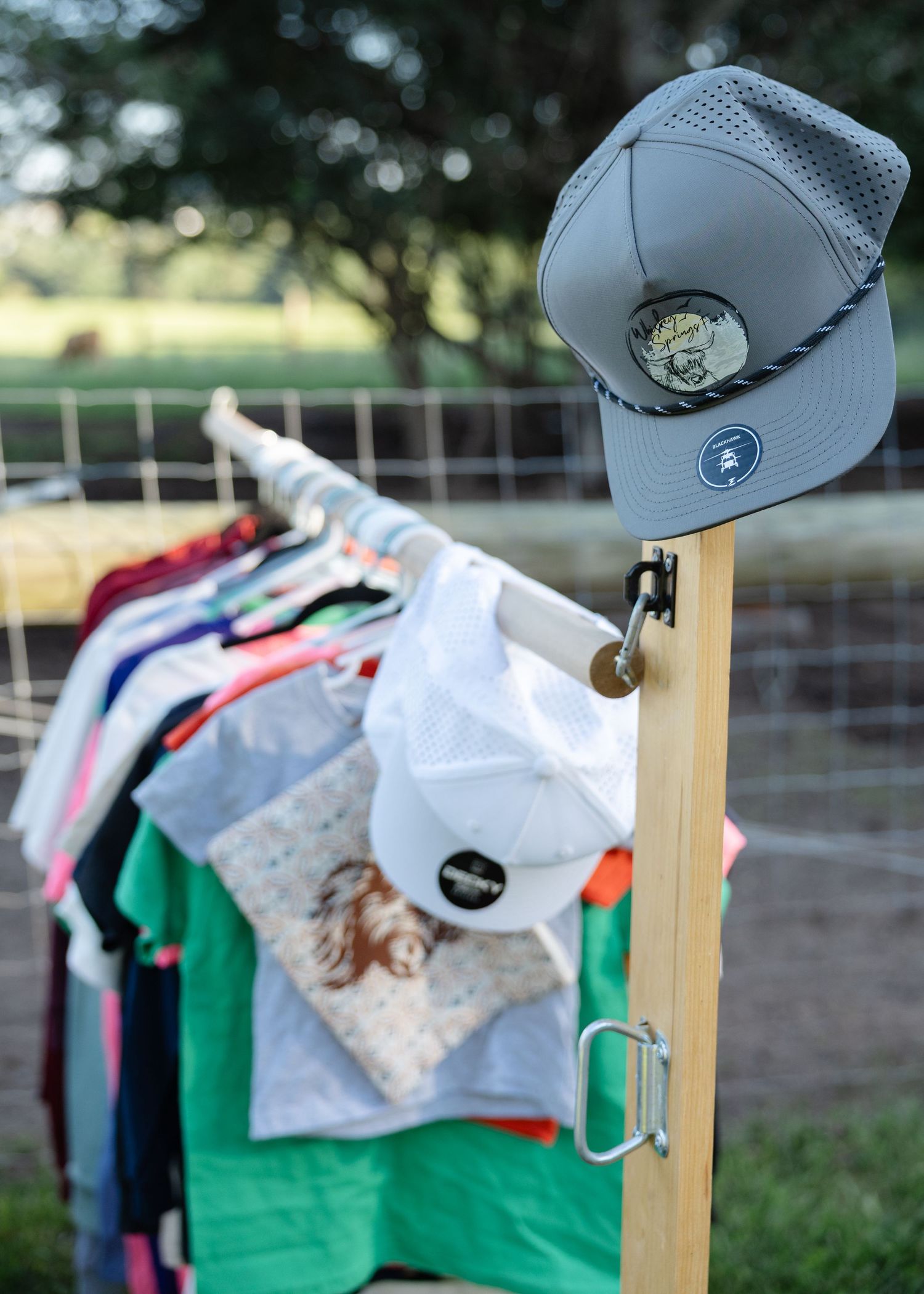 Outdoor clothing rack displaying colorful shirts and gray baseball caps near a farm fence.