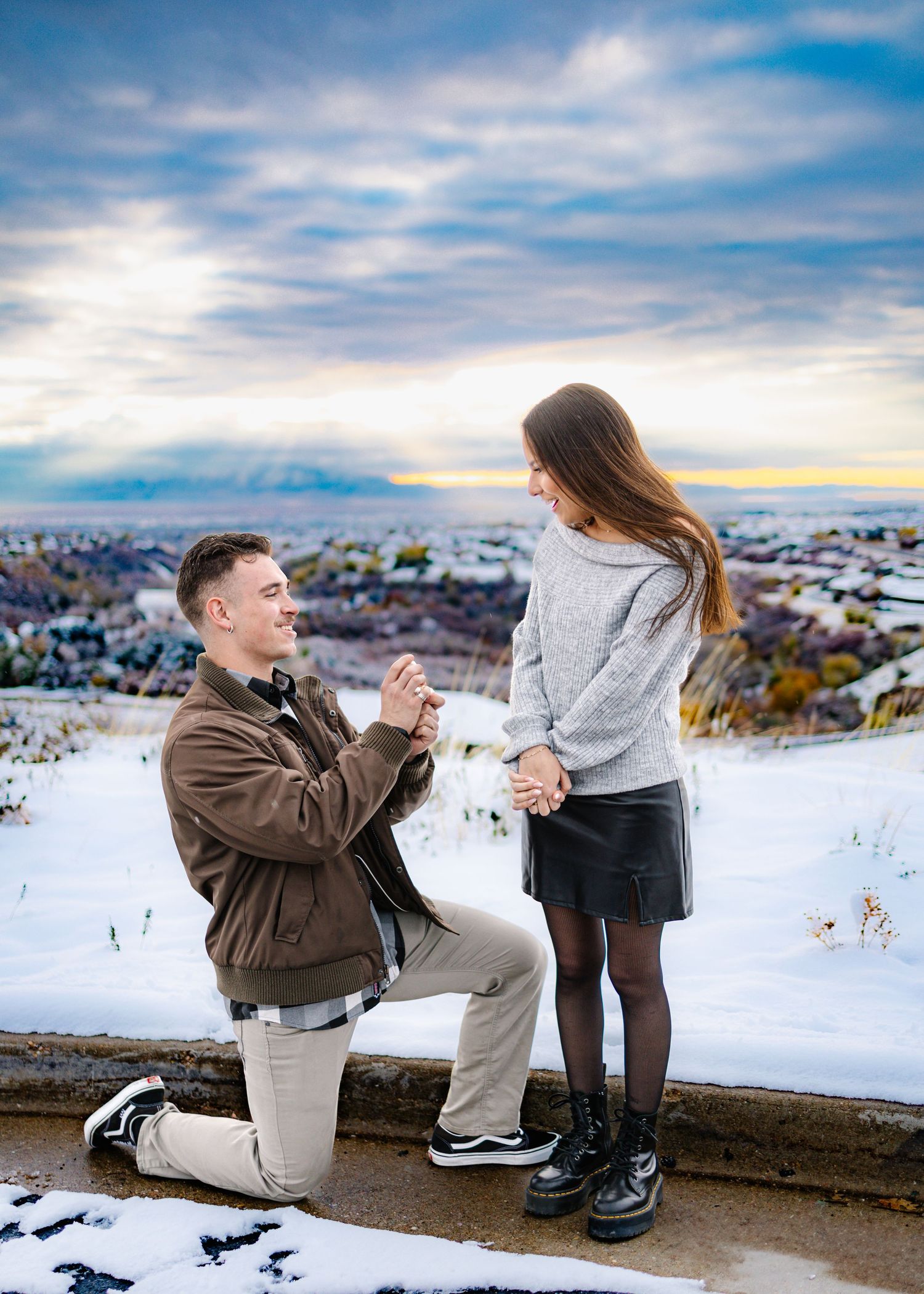 Man proposing on one knee to his girlfriend on a snowy overlook at a colorful winter sunset.