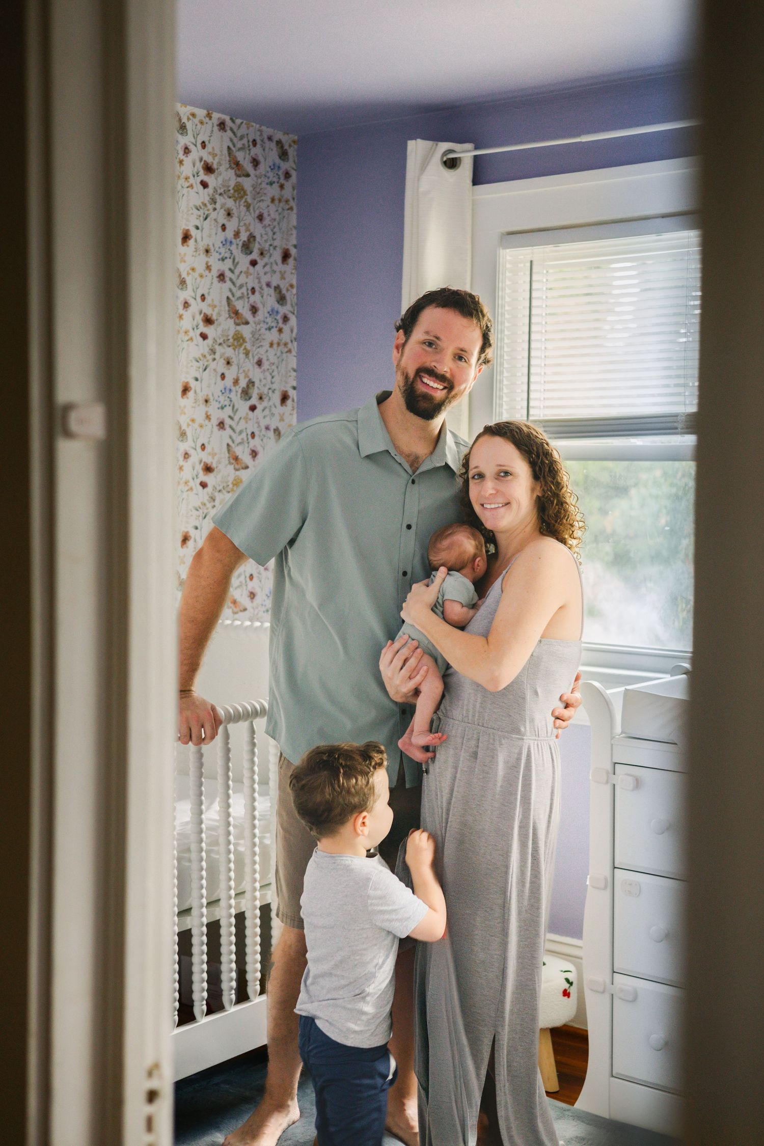 Happy family of four with toddler and newborn poses smiling in bright decorated baby nursery room.