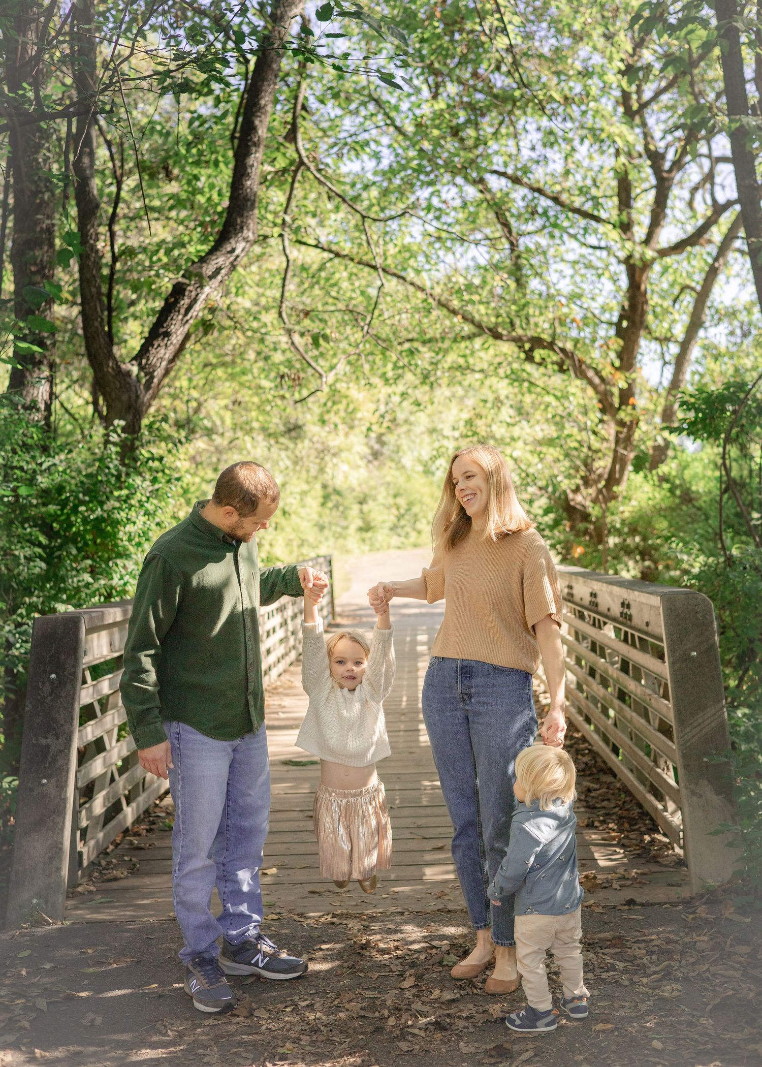 Family of four on a wooden bridge in a lush green forest, parents swinging toddler between them.