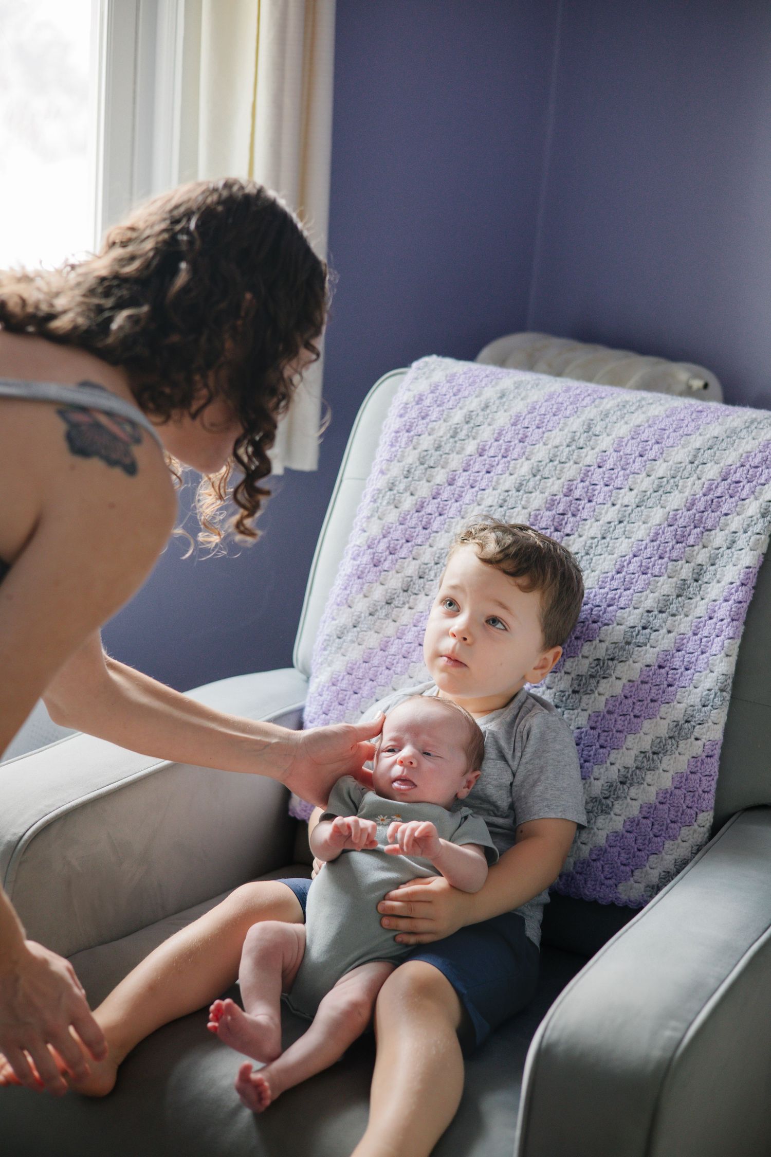 Mother carefully places newborn baby into arms of proud toddler boy sitting in nursery chair.