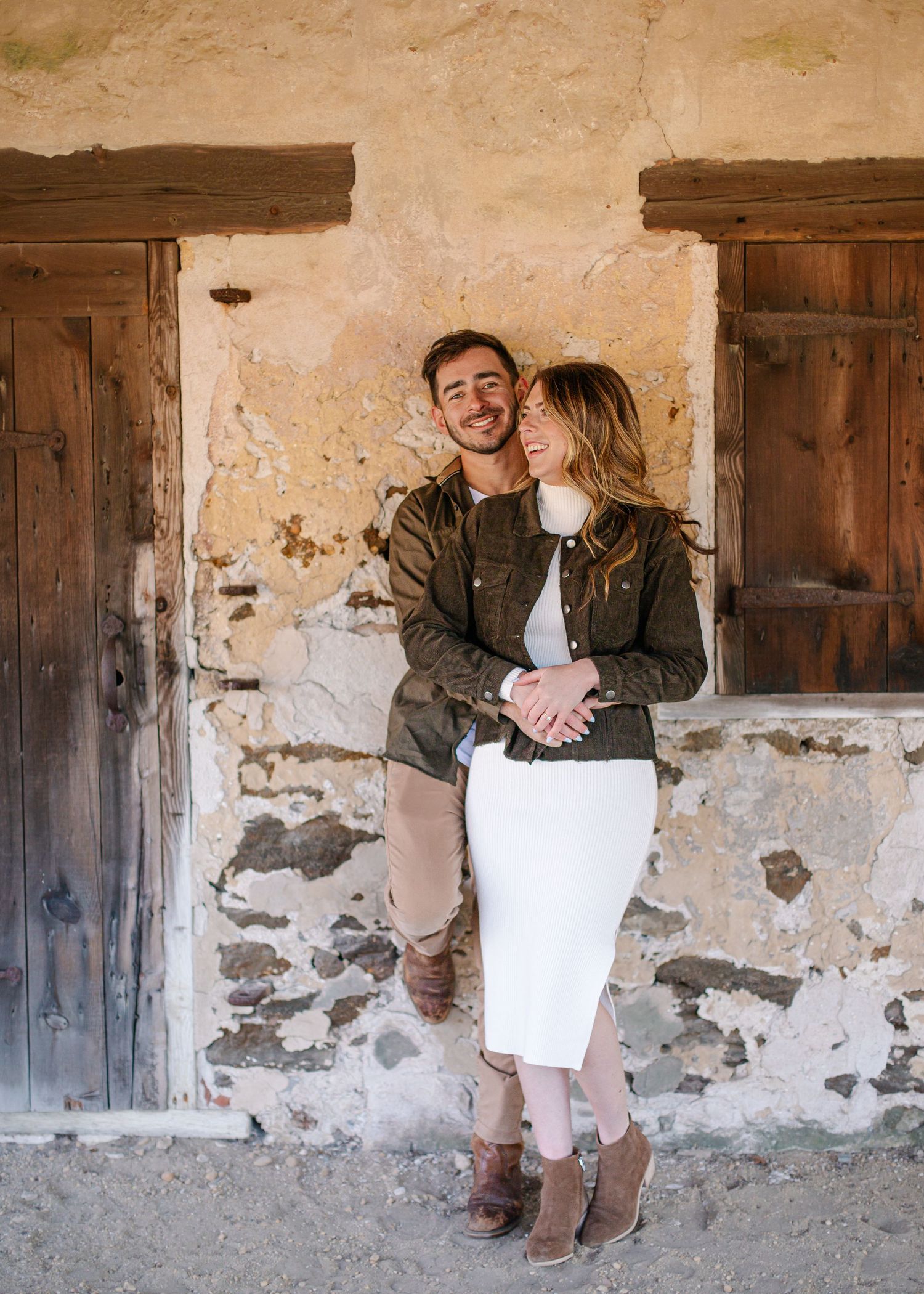 Smiling couple posing together against a rustic stone building with weathered wooden doors.