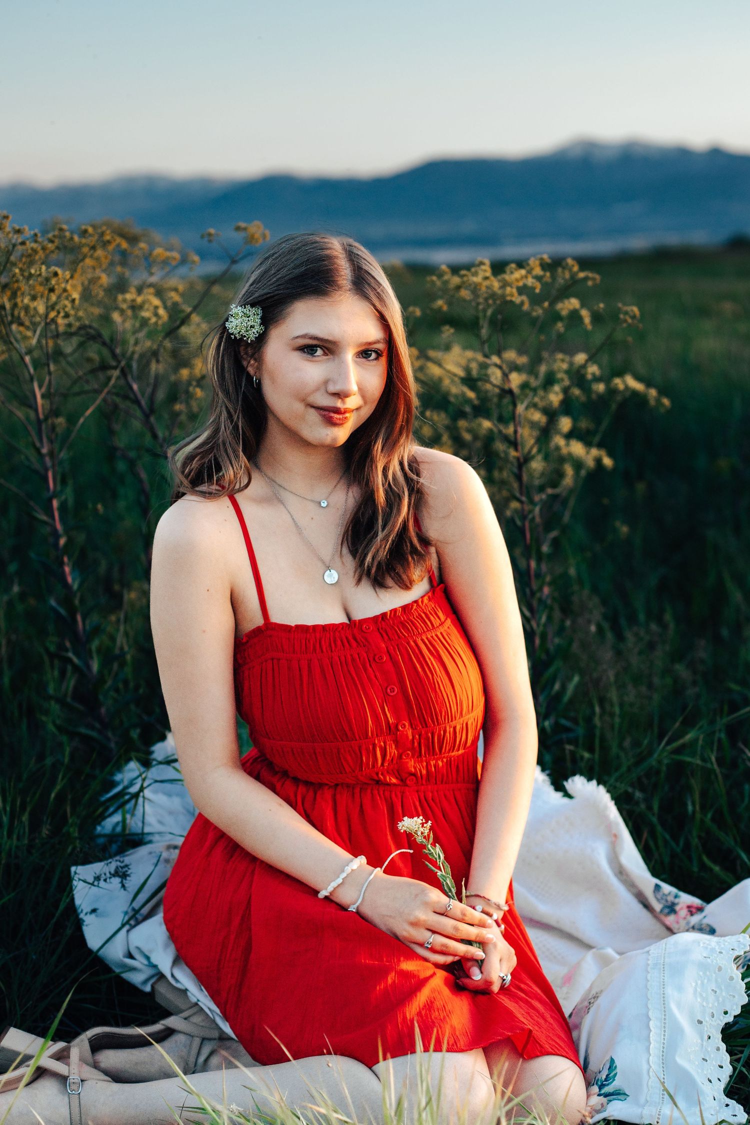 Young woman in a red dress sitting in a field at sunset with mountains in the background.