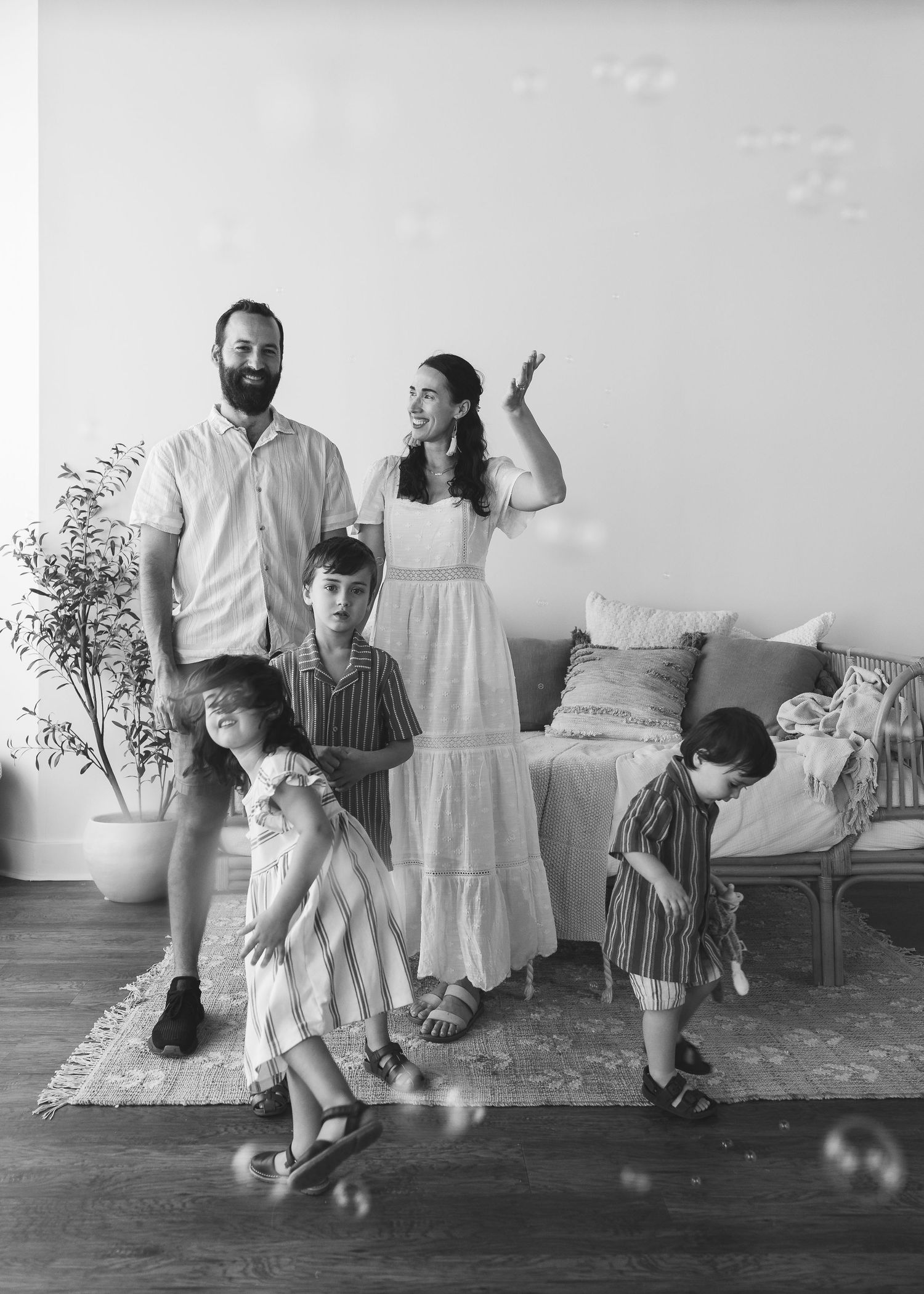 Black and white family portrait with parents and three children playing with bubbles in a bright living room.