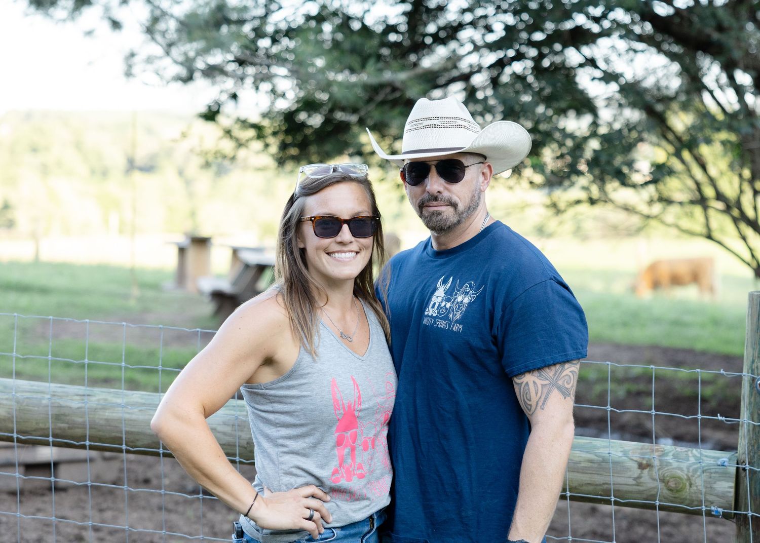 A couple wearing sunglasses poses outdoors near a fence, man in cowboy hat and blue tee, woman in gray tank top.