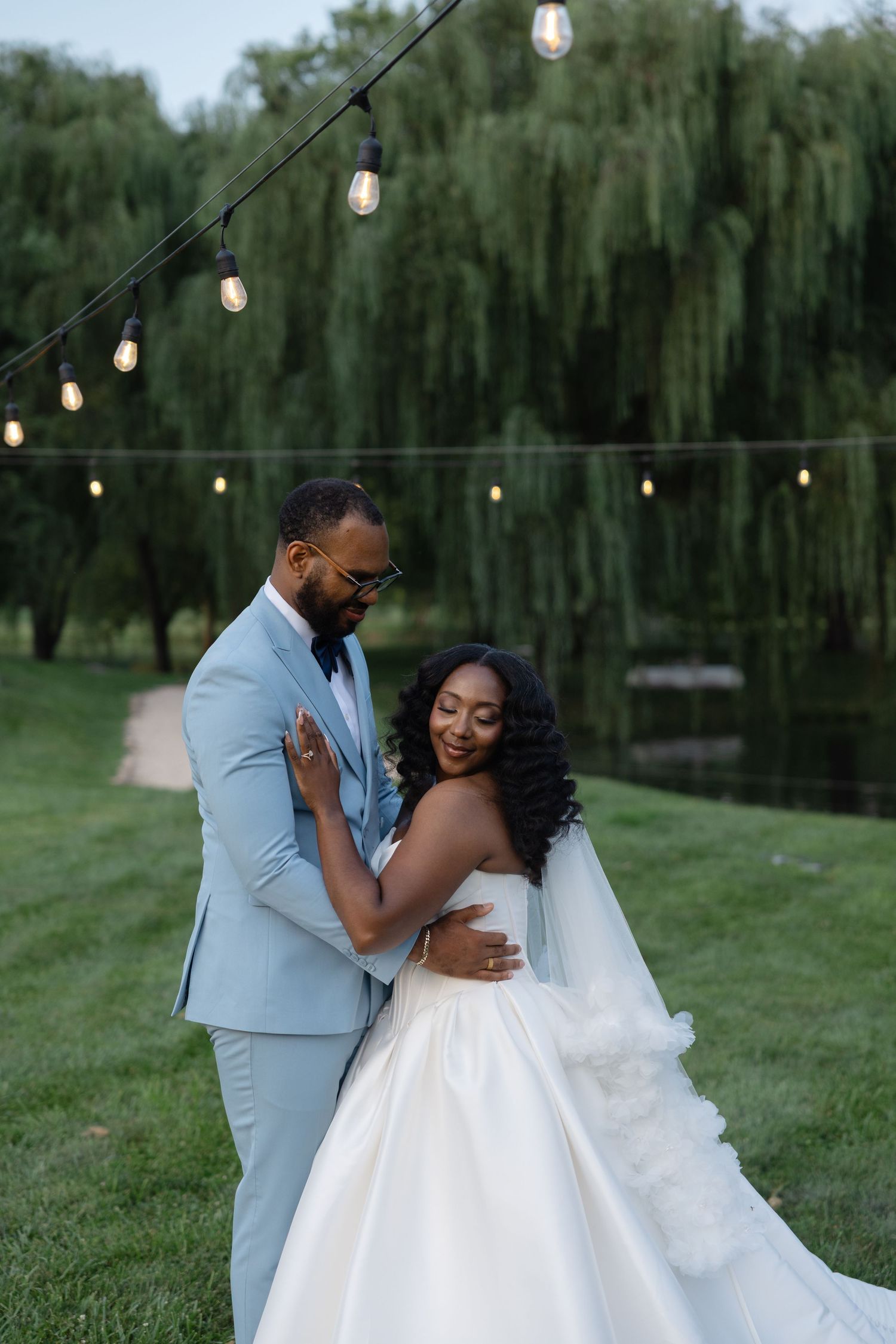 A bride and groom embrace outdoors under string lights with a weeping willow backdrop at dusk.