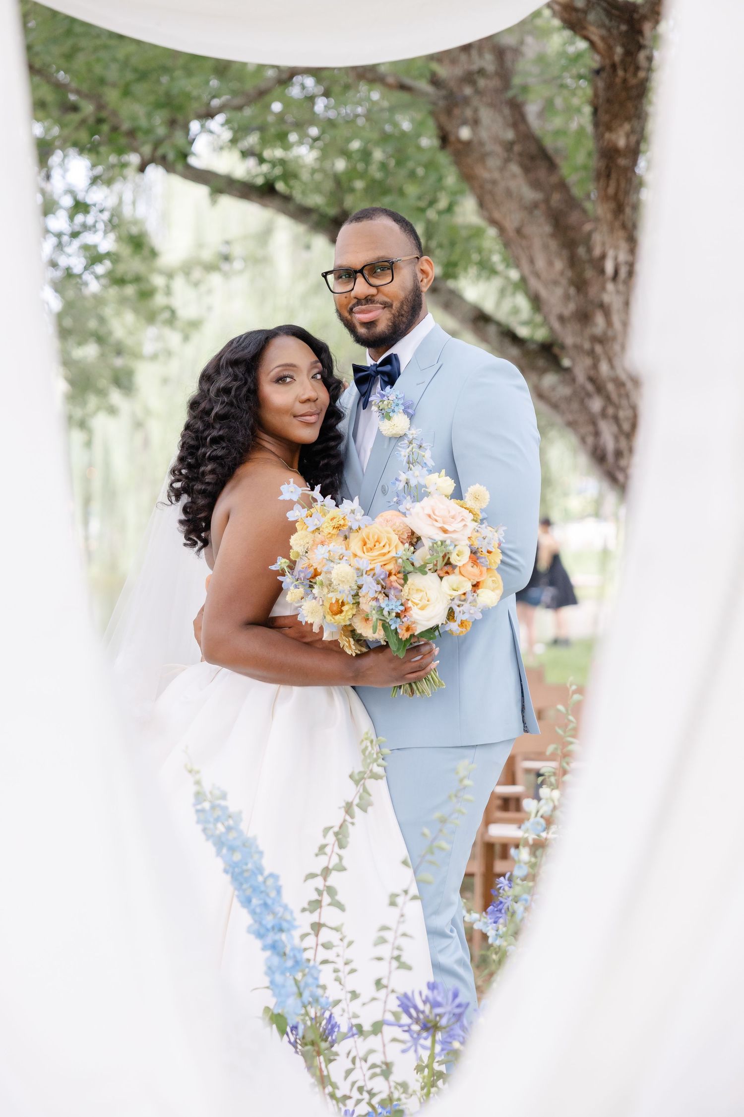 Bride and groom pose together framed by white draping with colorful floral bouquet outdoors.