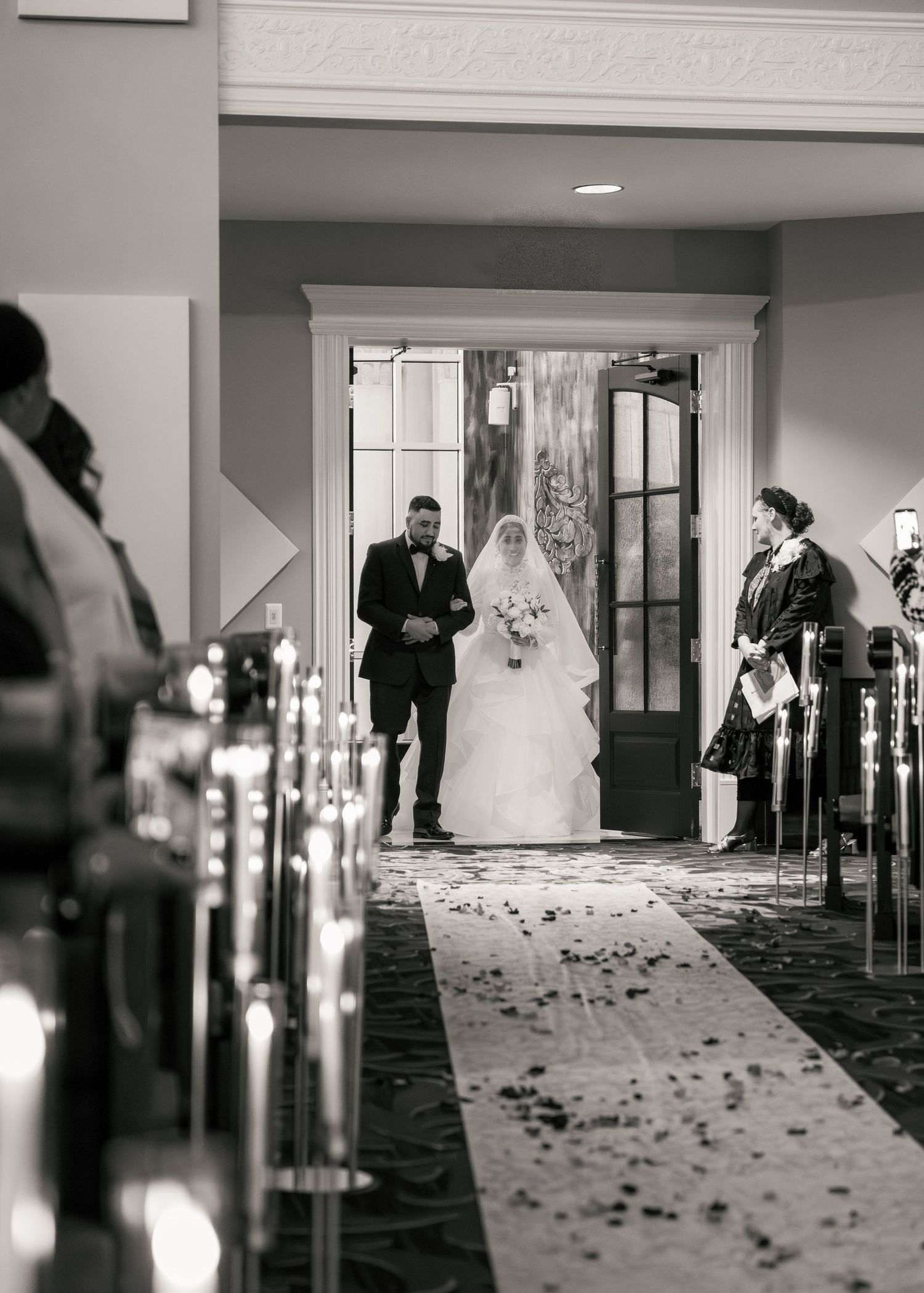 Bride walks down aisle with escort in black and white wedding ceremony photo.