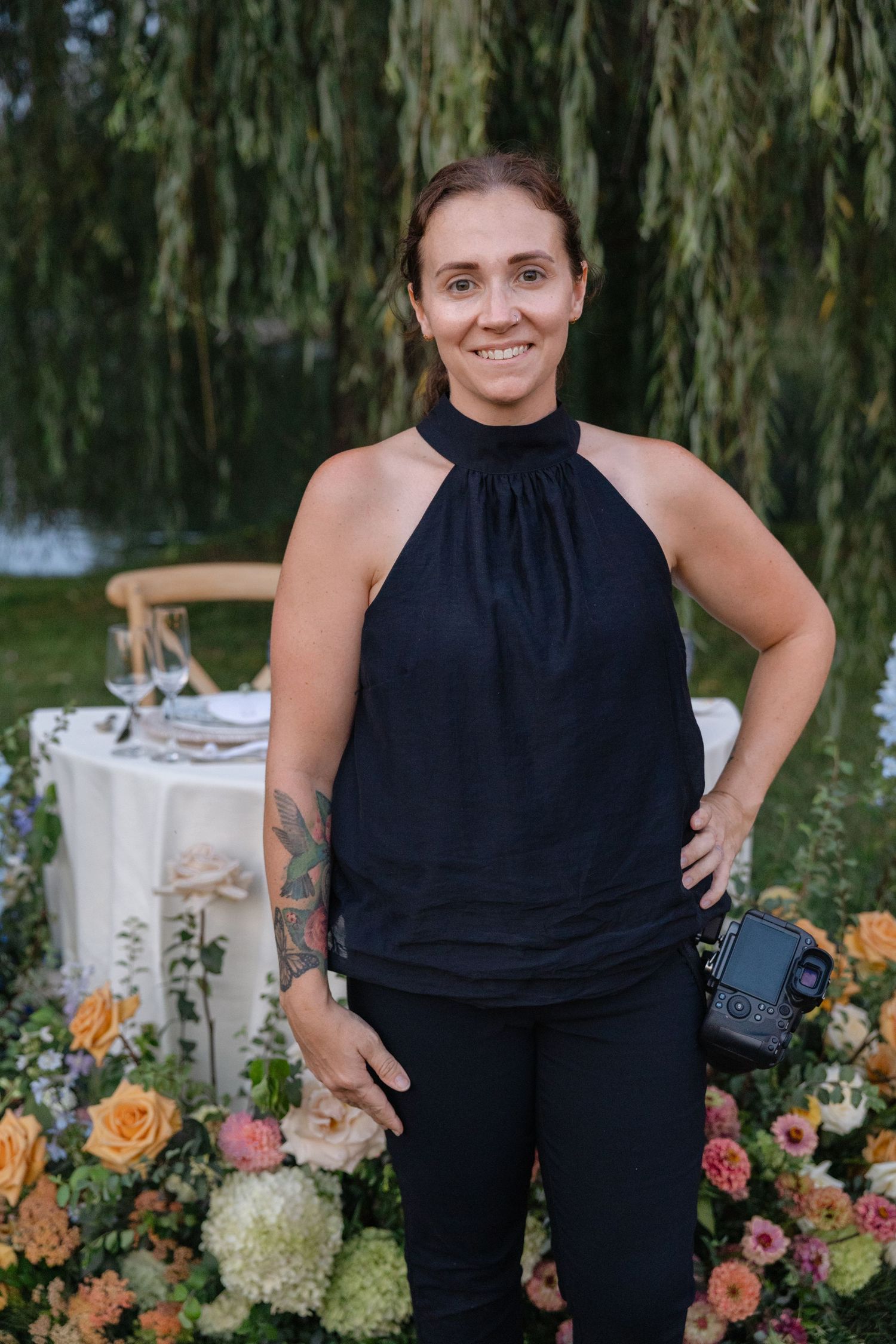 Female wedding photographer smiling in black halter top, standing near floral arrangements under a weeping willow tree.