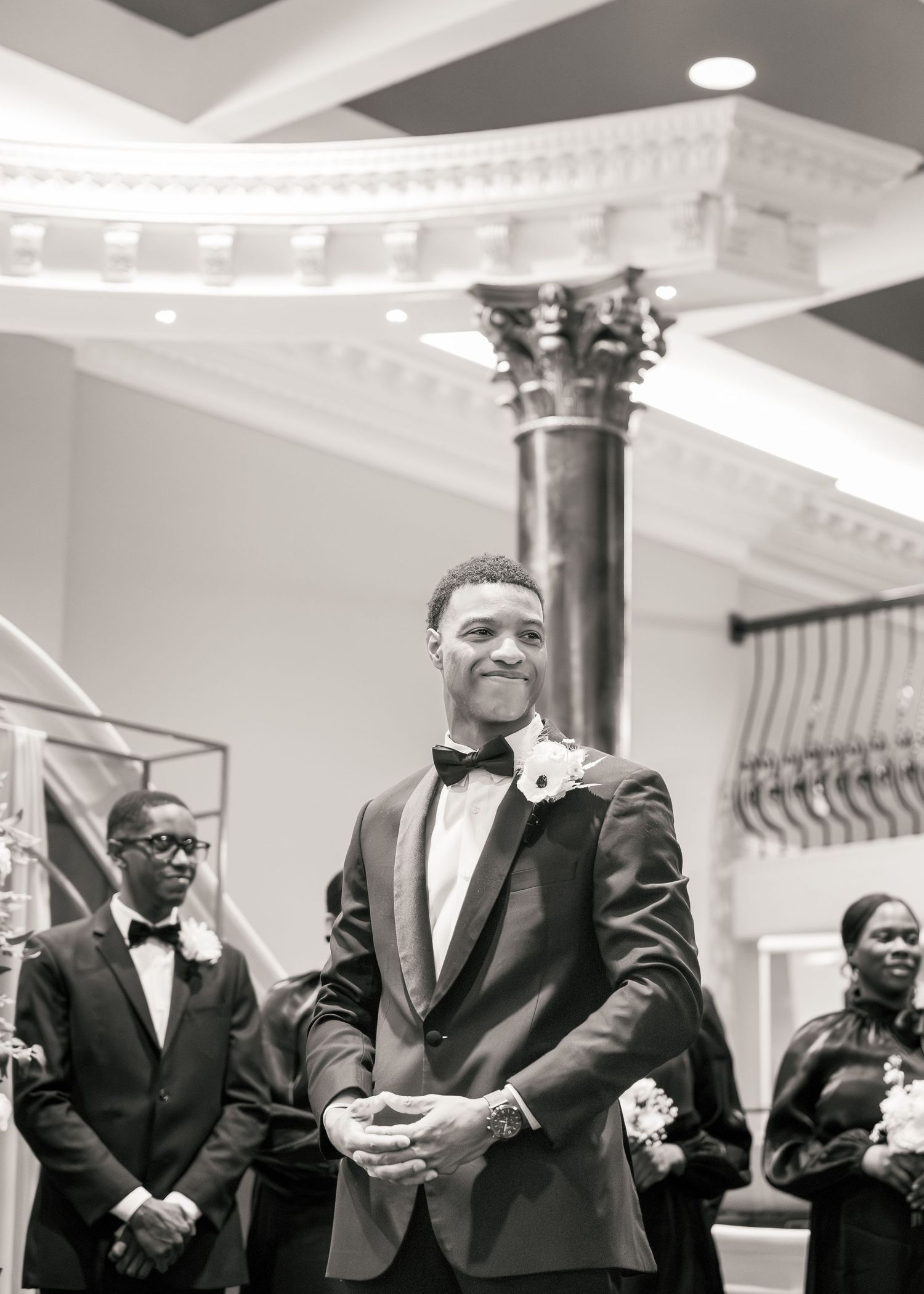 Groom in black tuxedo smiles at altar during elegant wedding ceremony in ornate venue.