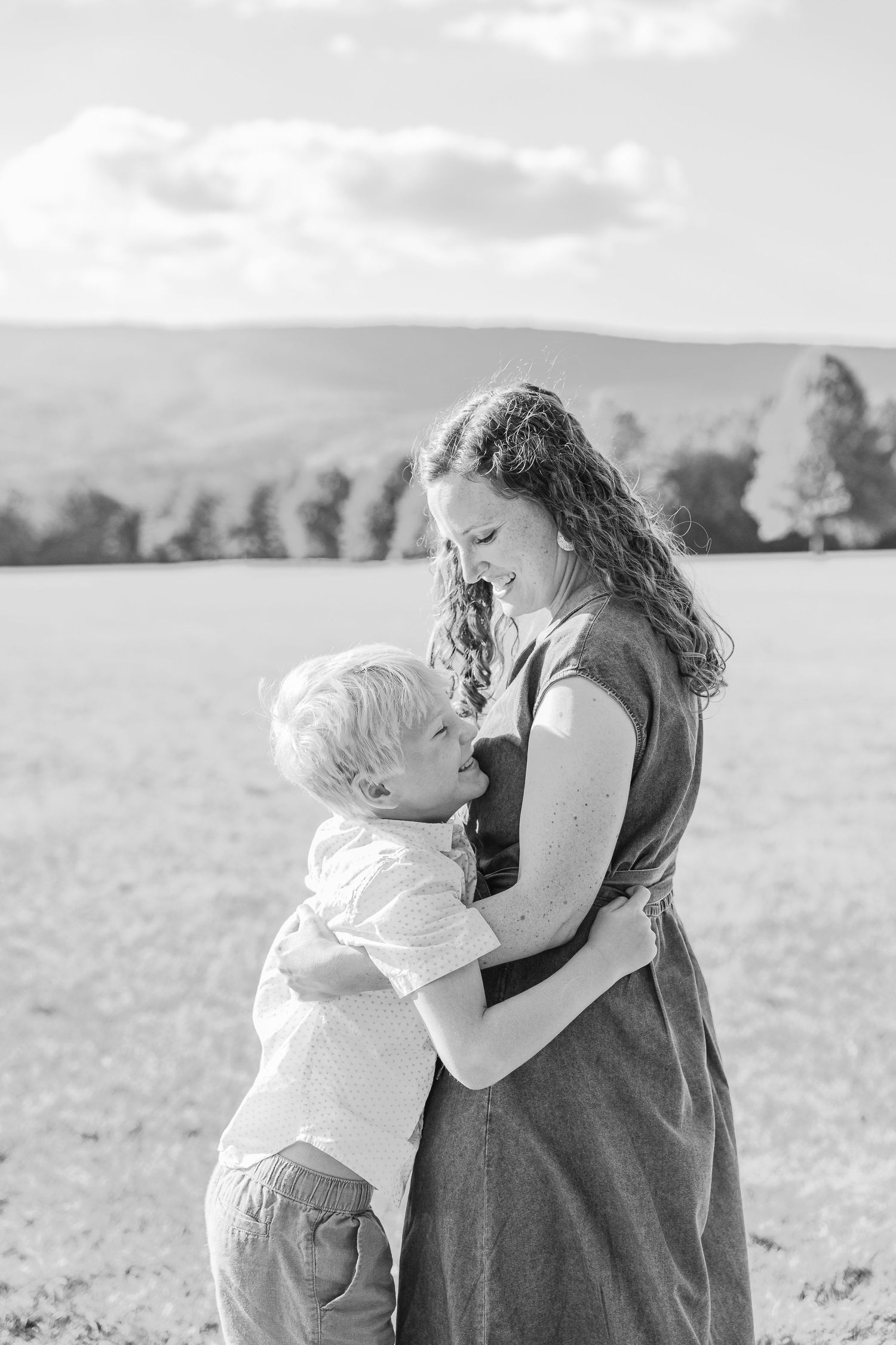 Black and white photo of a mother and young son sharing a warm embrace in an open field with rolling hills.