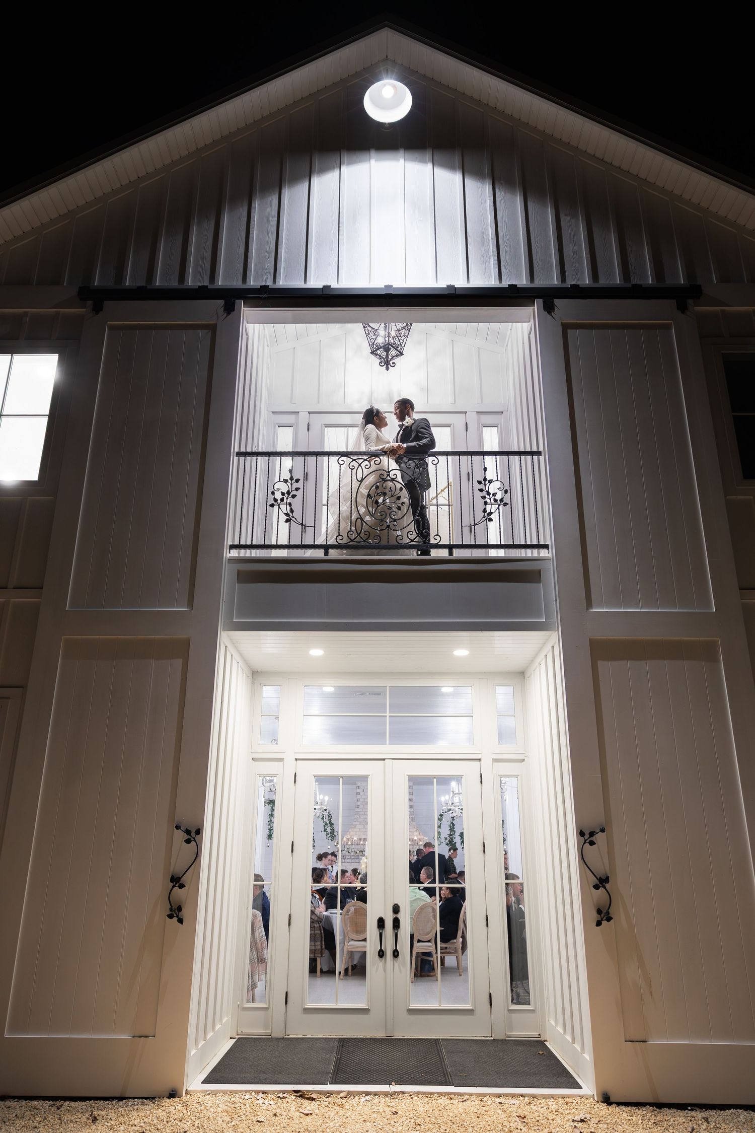 Bride and groom share a romantic kiss on a barn venue balcony at night with reception below.