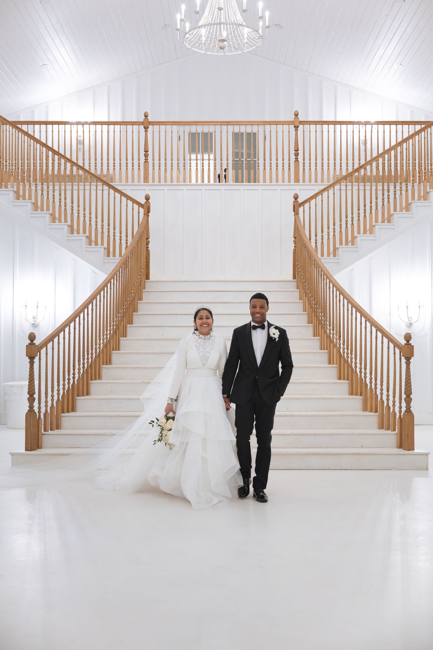 Bride and groom holding hands walking down grand white staircase in elegant wedding venue.
