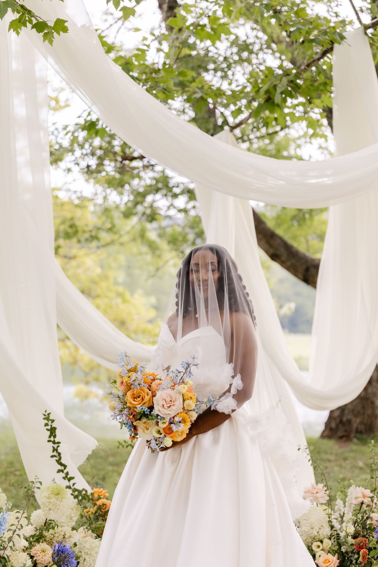 Bride wearing a white gown and veil holding a colorful bouquet under white draped fabric outdoors.