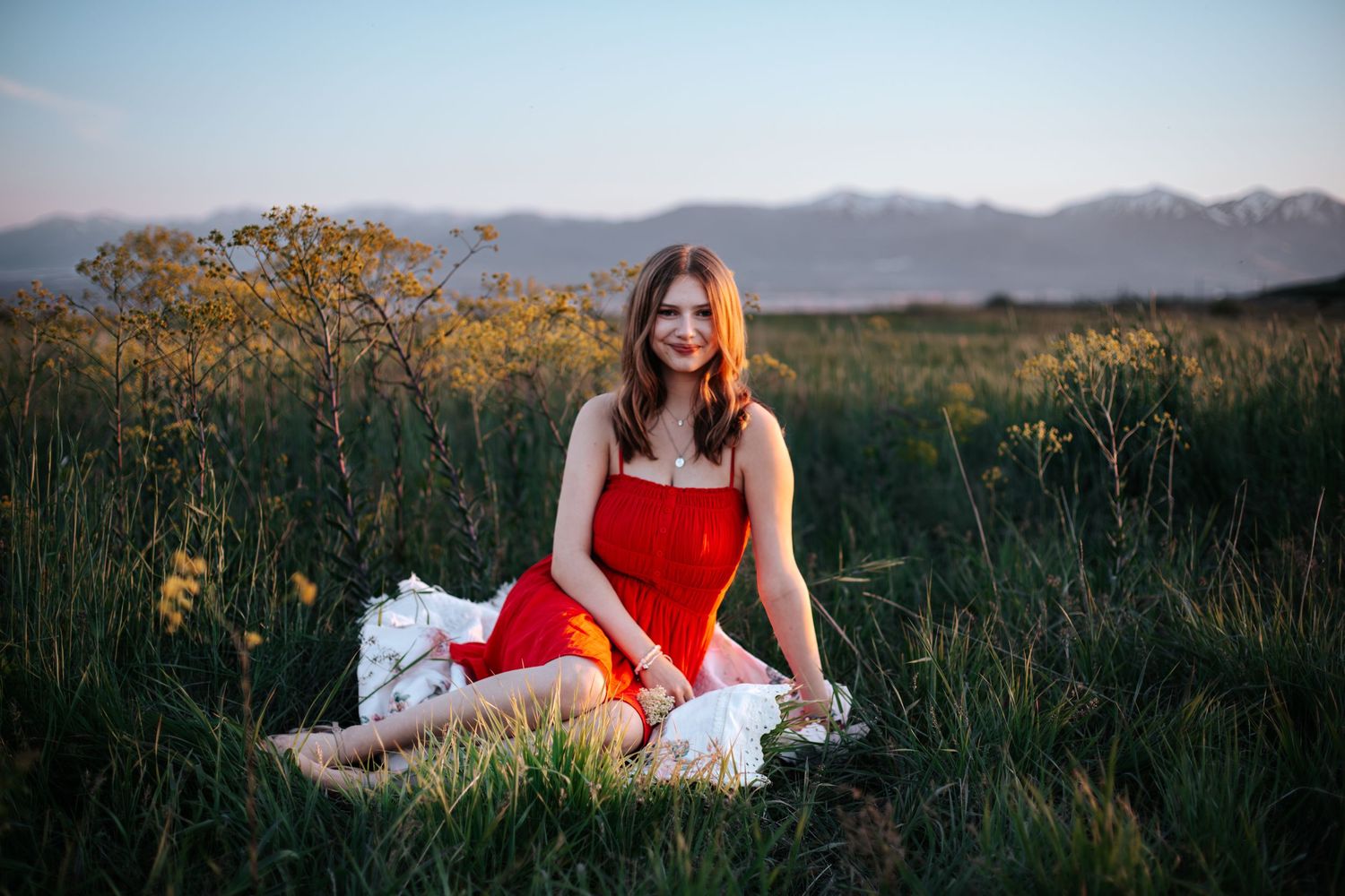 Young woman in red dress sitting on a blanket in a green meadow with mountains and golden sunset in background.