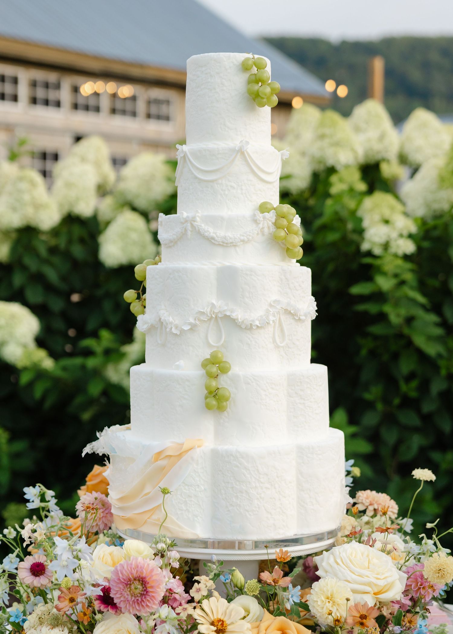 Elegant five-tier white wedding cake decorated with green grapes and draped fondant, surrounded by colorful flowers.
