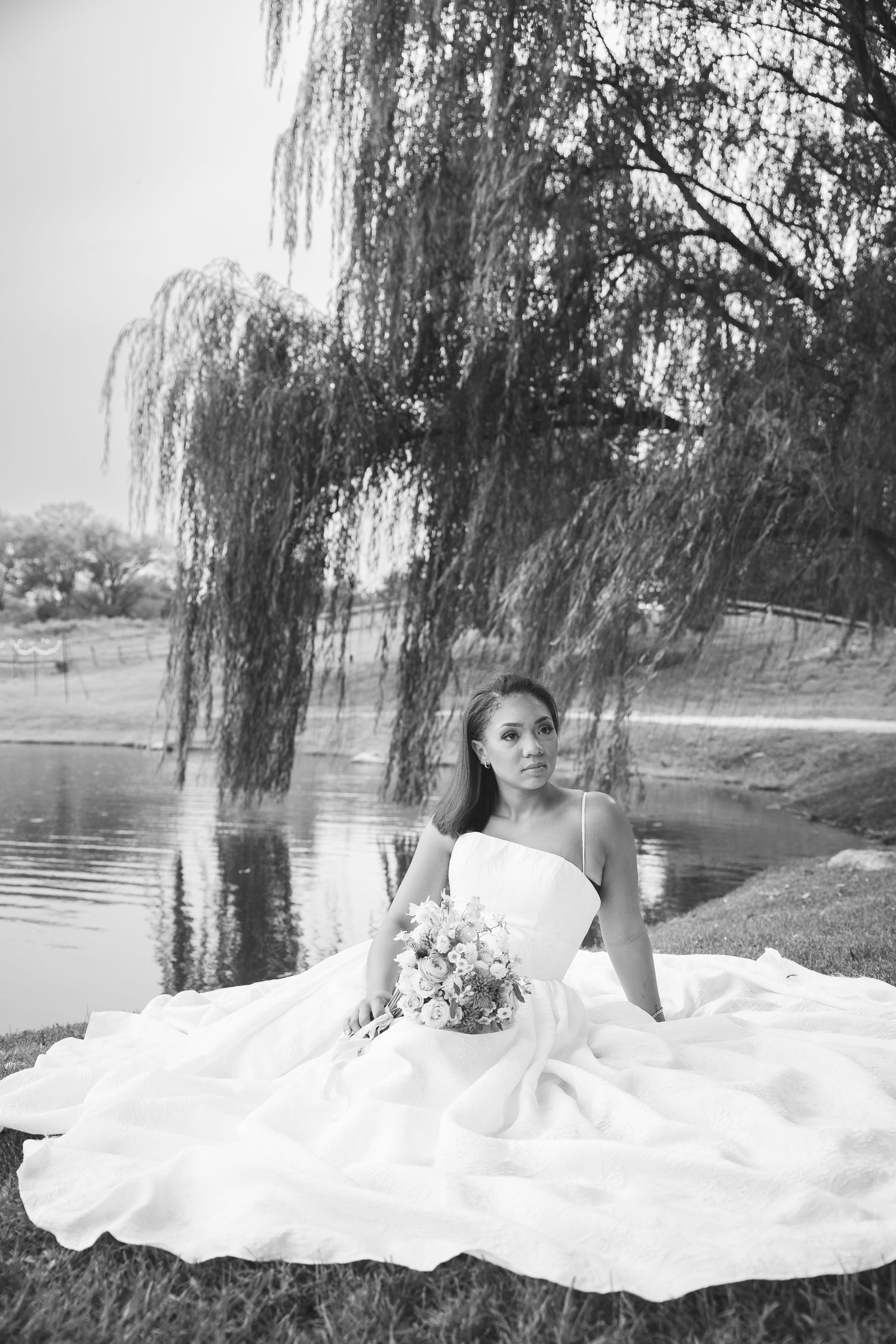 Black and white portrait of bride in strapless gown seated by a pond under a weeping willow holding a bouquet.