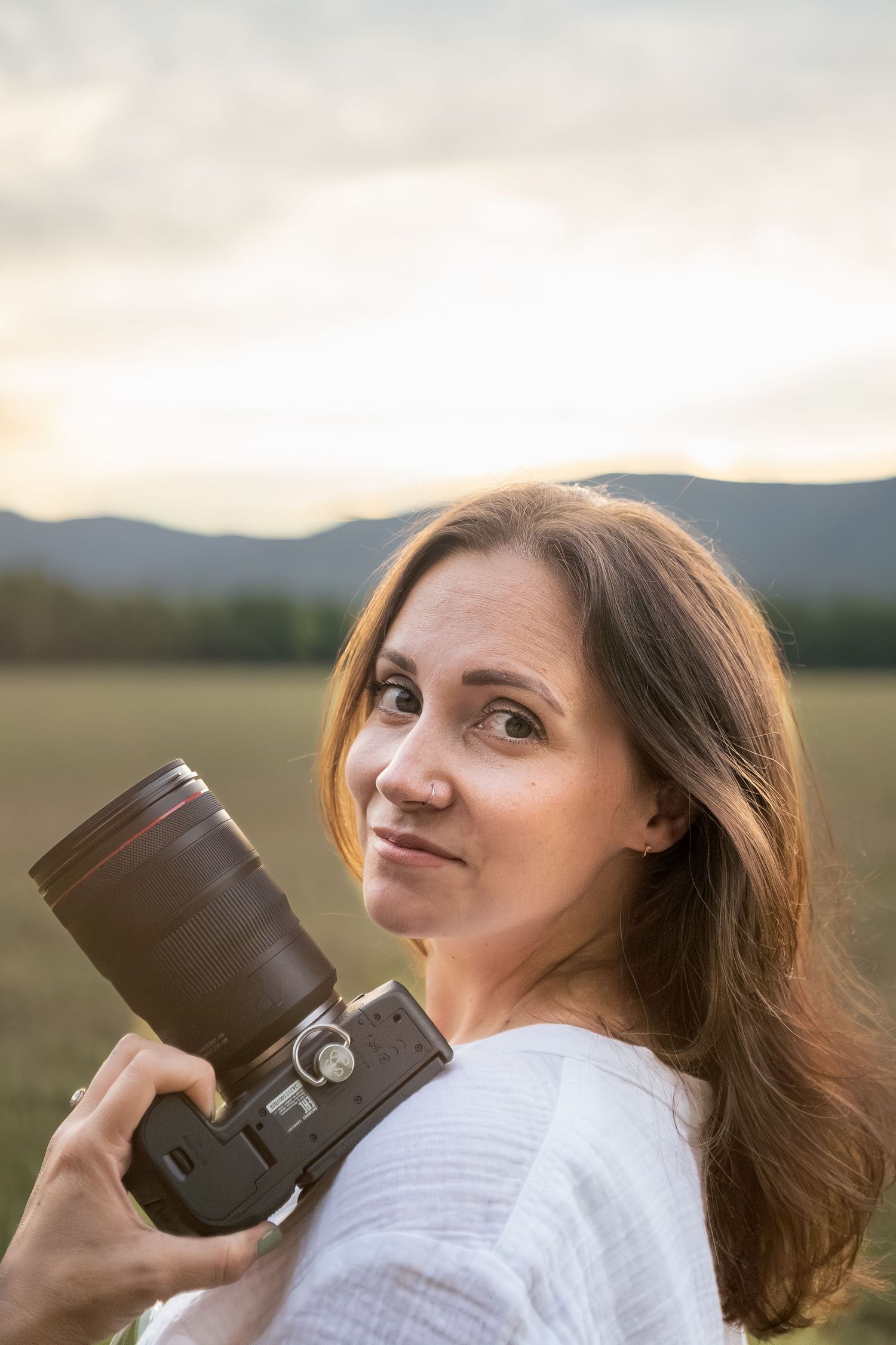 Woman photographer holding DSLR camera outdoors at sunset with mountains and field in background.