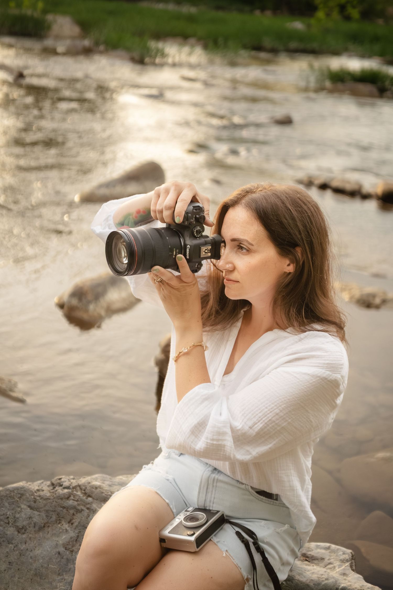 Female photographer sitting on rocks by a river, focusing her DSLR camera during golden hour.