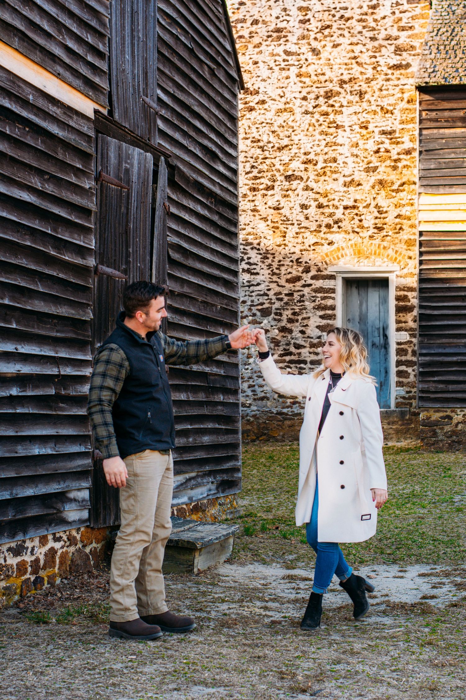 Couple reaching out to hold hands between a dark wooden barn and a golden stone building.