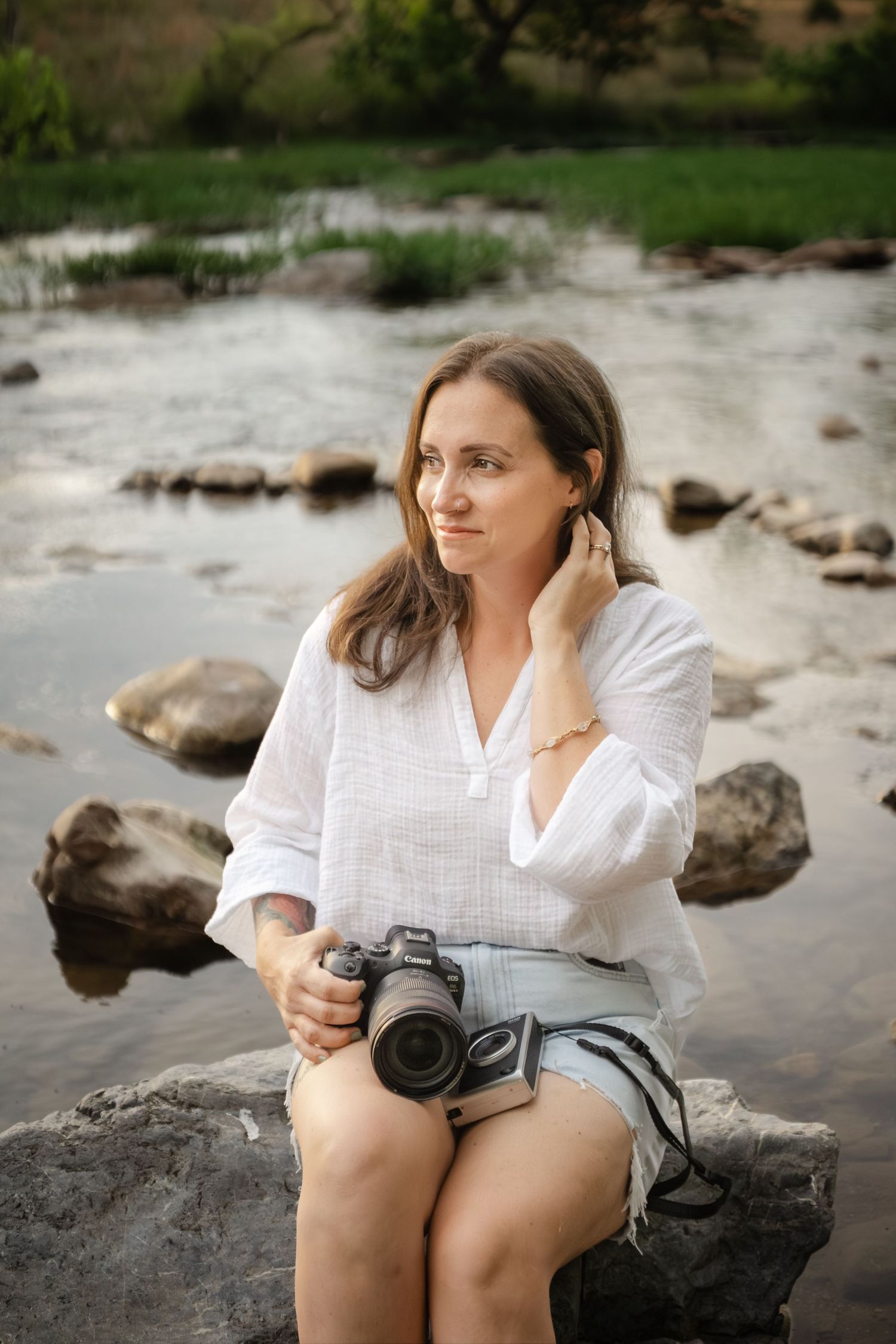 Woman photographer sitting on rocks by a stream, holding a DSLR camera, wearing white shirt and denim shorts.