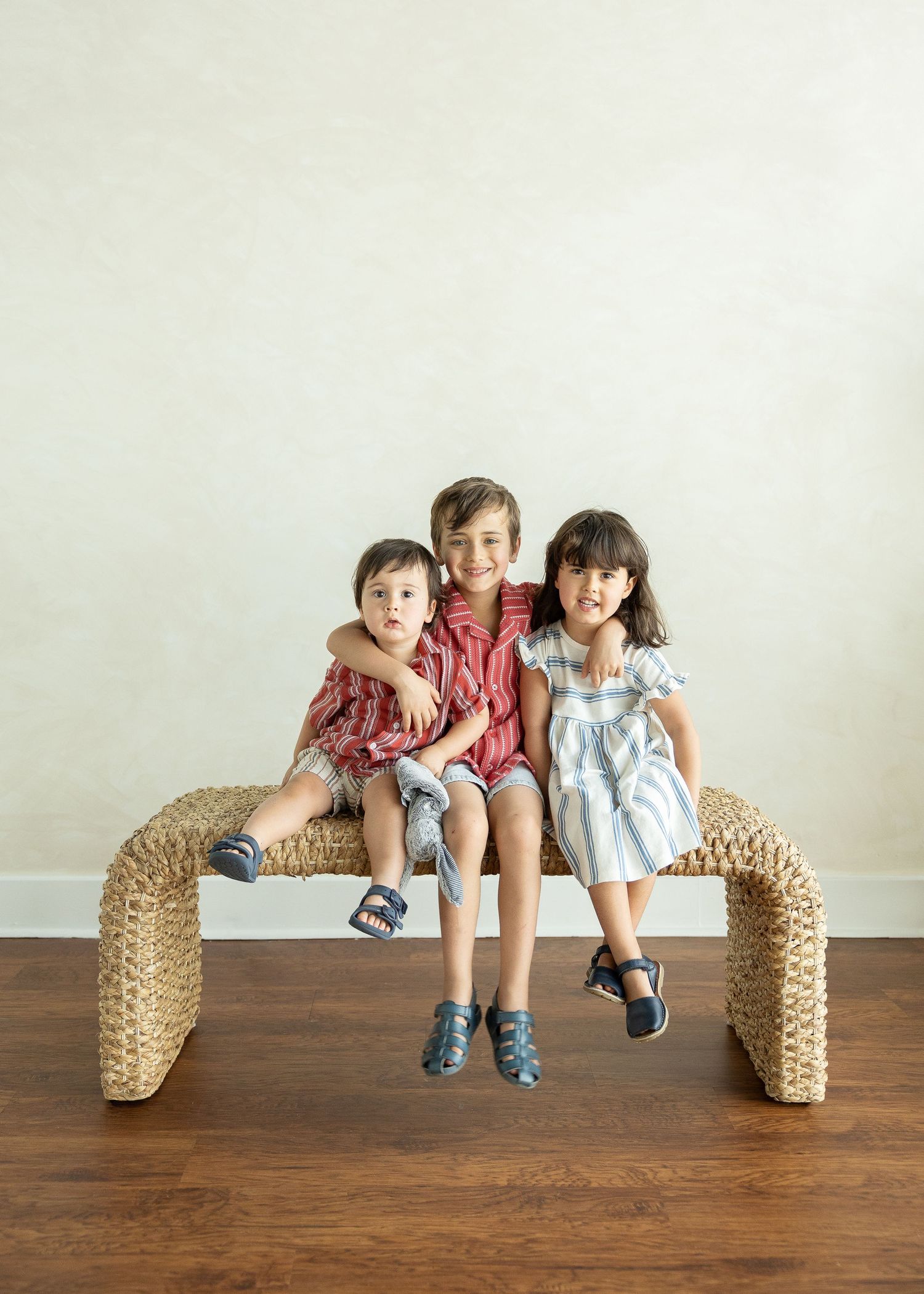 Three siblings sitting together on a woven bench, smiling at the camera in a bright studio.