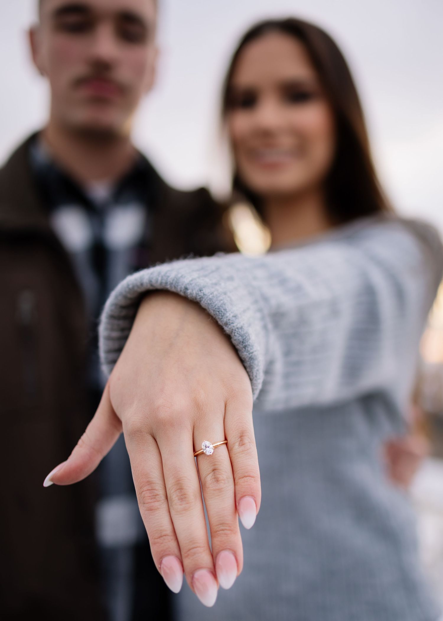 Engaged couple showing diamond ring close-up with blurred couple smiling in background.