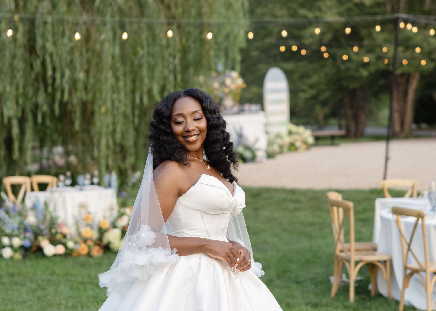 Smiling Black bride in strapless white ballgown with veil twirls at outdoor garden wedding reception.
