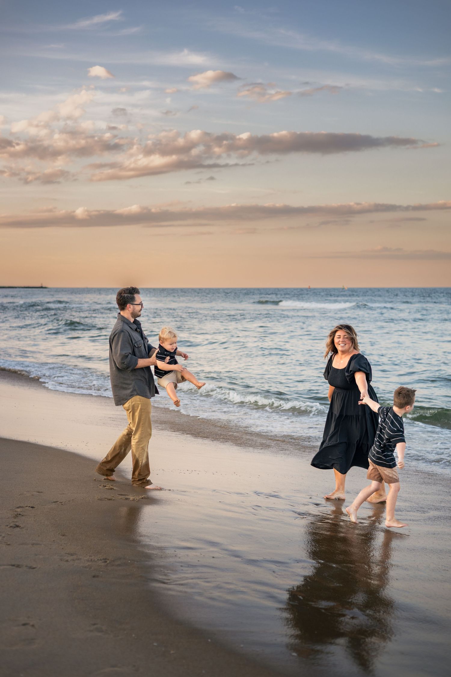 Family of four playing on a sandy beach at sunset, with parents swinging children near ocean waves.