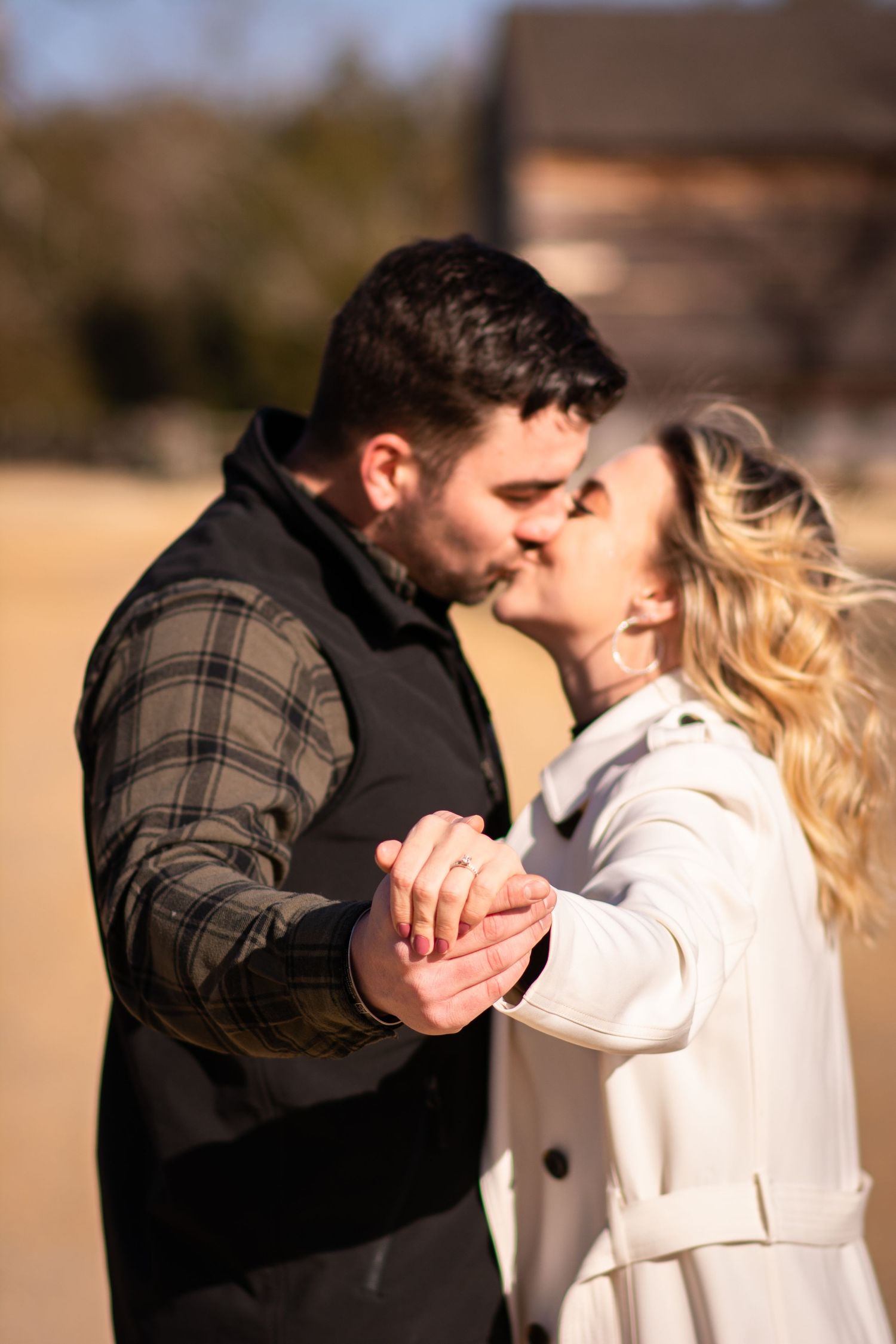 Couple sharing a kiss while holding hands outdoors, showing an engagement ring in warm sunlight.
